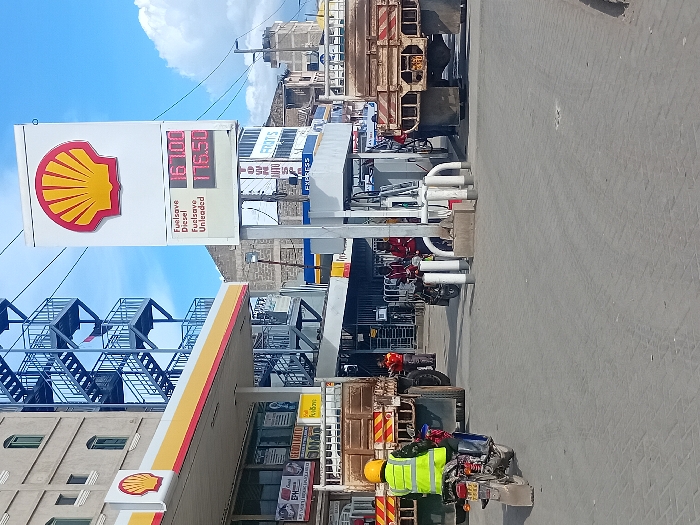 The scene takes place at a Shell gas station in Mlolongo, Kenya, located at coordinates -1.394 latitude, 36.94 longitude. The gas station is a busy one, with several vehicles present. A man in a yellow safety vest is on a motorcycle at the far left of the image, while two men are filling up their vehicles with gas at the pumps. Two trucks are parked to the right, blocking the view of the main road. The gas station sign displays the price of fuel: 167.00 for fuel-save diesel and 176.50 for fuel-save unleaded. The exterior of the building is painted in Shell's signature red and yellow colors. The gas station is in a bustling urban area, with buildings and other businesses in the background. The sky is blue with a few white clouds.