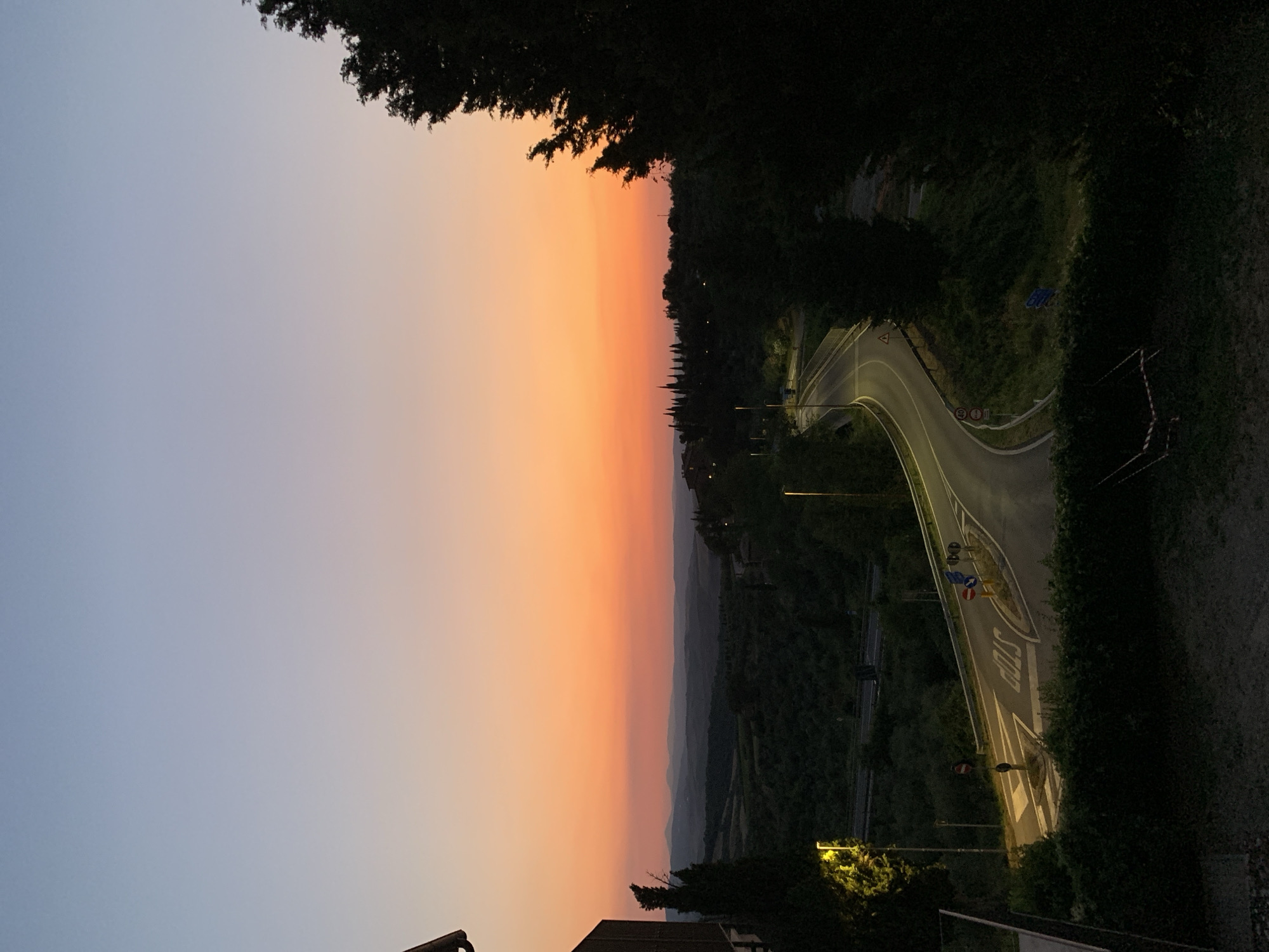The image captures a picturesque Tuscan landscape at sunset, viewed from an elevated position overlooking a winding road. The sky is a gradient of soft blues and warm oranges, indicative of dusk. Rolling hills, cloaked in lush greenery and dotted with sparse buildings, recede into the distance, shrouded in a gentle haze. A tree-lined road curves through the scene, illuminated by streetlights and reflecting the fading sunlight. Several road signs are visible, including what appears to be a "STOP" sign painted on the asphalt and other traffic signs. The overall mood is serene and tranquil, evoking the beauty of an Italian countryside evening.