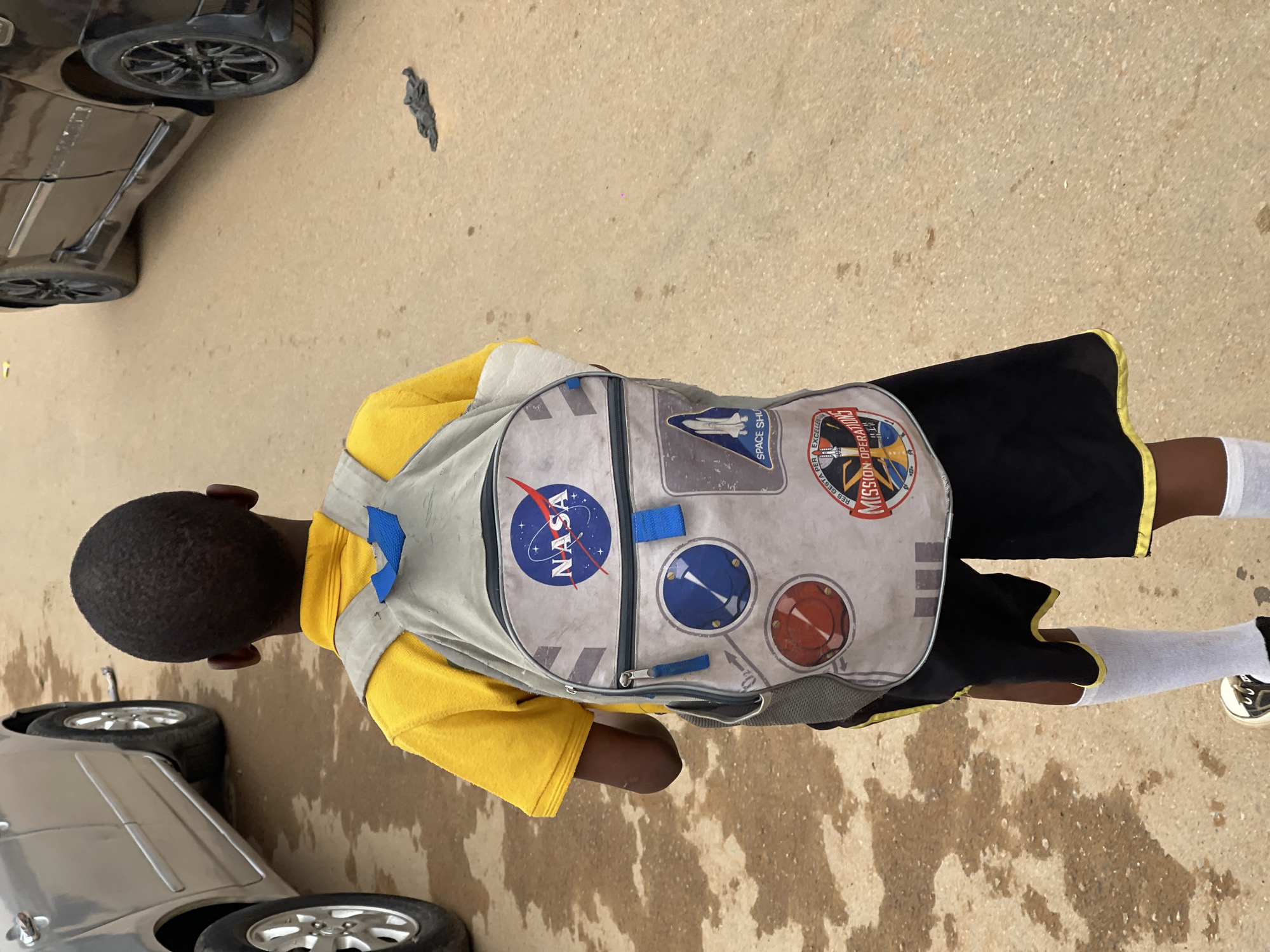 A young boy stands on a paved surface, presumably a street, in Karmajiji, Nigeria, with a car visible in the lower left and upper left corners. He is wearing a yellow t-shirt, black shorts with a yellow trim, and white socks. He has a NASA-themed backpack. The background features a ground covered in a substance that appears to be water, though it could be stains on the pavement.