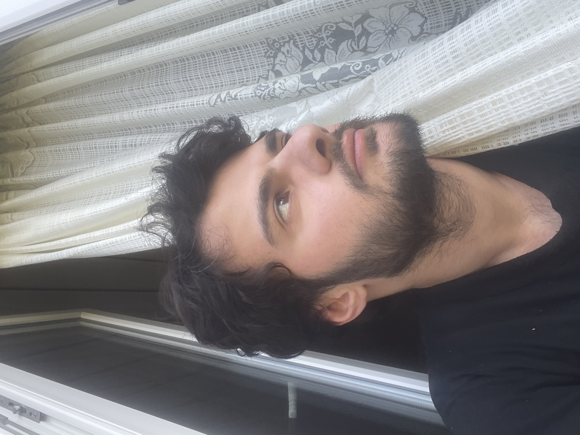 A young man with dark, curly hair and a beard is looking out of a window, partially obscured by sheer white curtains with a floral pattern. He is wearing a black t-shirt. The window frame is visible to the left of the frame, with a handle on the outside. The scene appears to be indoors, with soft natural light filtering through the curtains. The background suggests an urban environment.