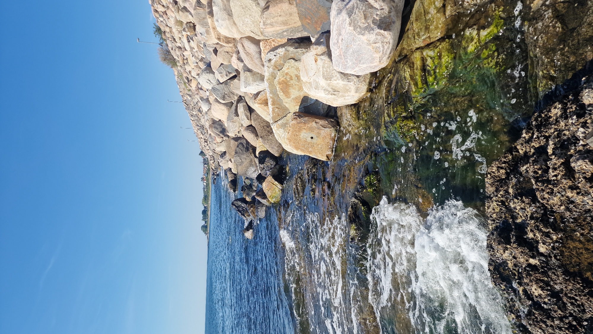 The image depicts a rocky coastline in Simrishamn, Sweden, under a clear blue sky. The foreground features a large, dark, rough-textured rock with waves gently lapping at its base. The water is a deep blue, transitioning to a clearer, shallower area where waves are breaking over submerged rocks covered in green algae. A line of large, rounded boulders forms a breakwater extending along the coastline to the right. In the distance, a town with houses and trees is visible along the shore, with a few streetlights dotting the breakwater. The overall scene is a tranquil coastal landscape.