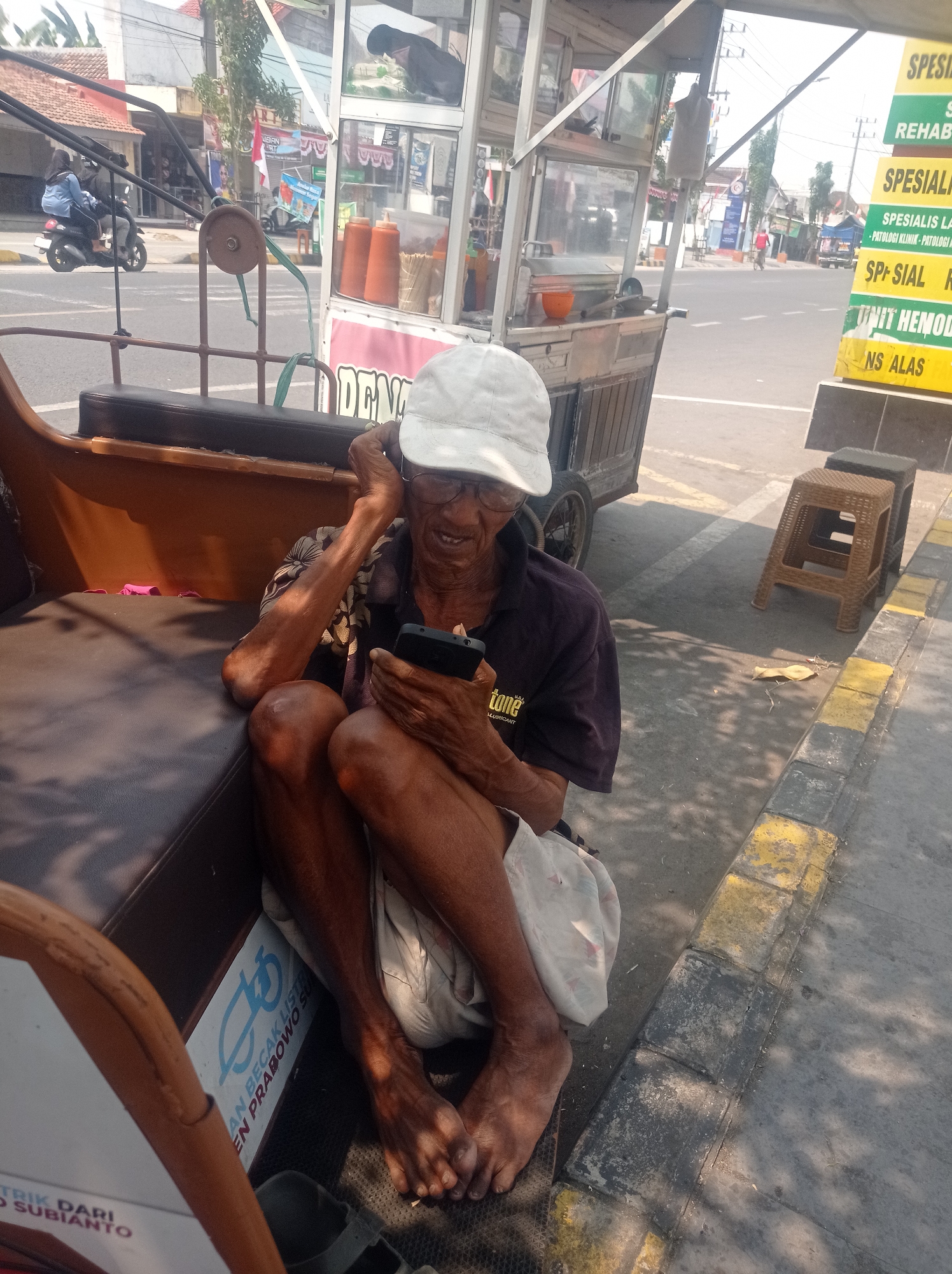 The image depicts an elderly Indonesian man sitting on the edge of what appears to be a pedicab in Bangunsari, Indonesia. He is wearing a white cap and glasses, and is holding a smartphone in his hands. The man is seated next to a food cart, which is typical of street vendors in the area. In the background, there are buildings and a road with traffic, suggesting a busy urban environment. The lighting indicates it is daytime, with sunlight casting shadows on the ground. The overall scene captures a moment of everyday life in a bustling Indonesian city.