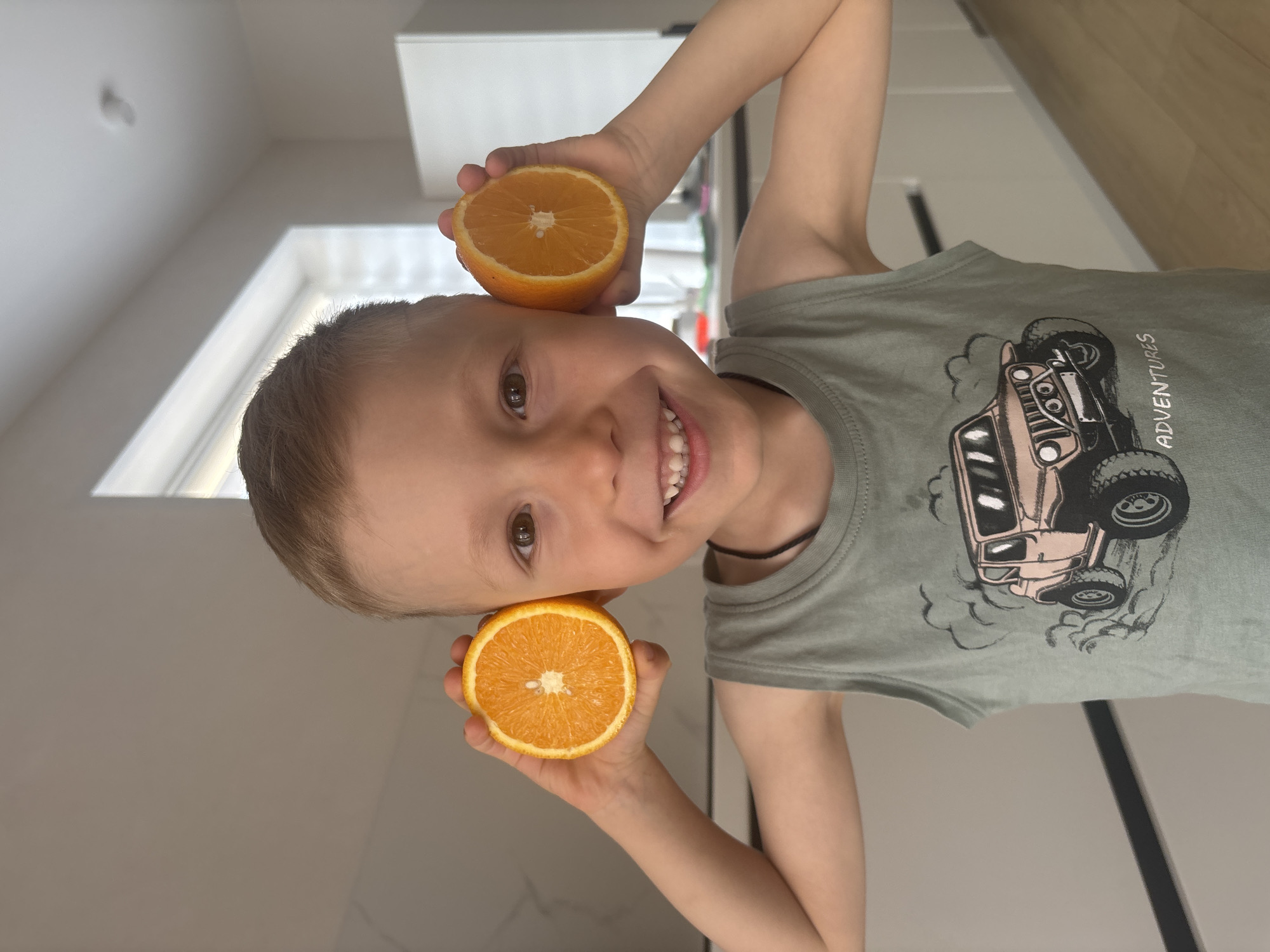 A smiling young boy is holding two halves of an orange up to his ears, appearing as though they are earphones or earmuffs. He is wearing a light green tank top with a cartoon truck graphic on it. The background is a modern kitchen with white cabinets and a light-colored countertop or wall. The location might be Chaikovsky, Russia, as specified in the instructions.