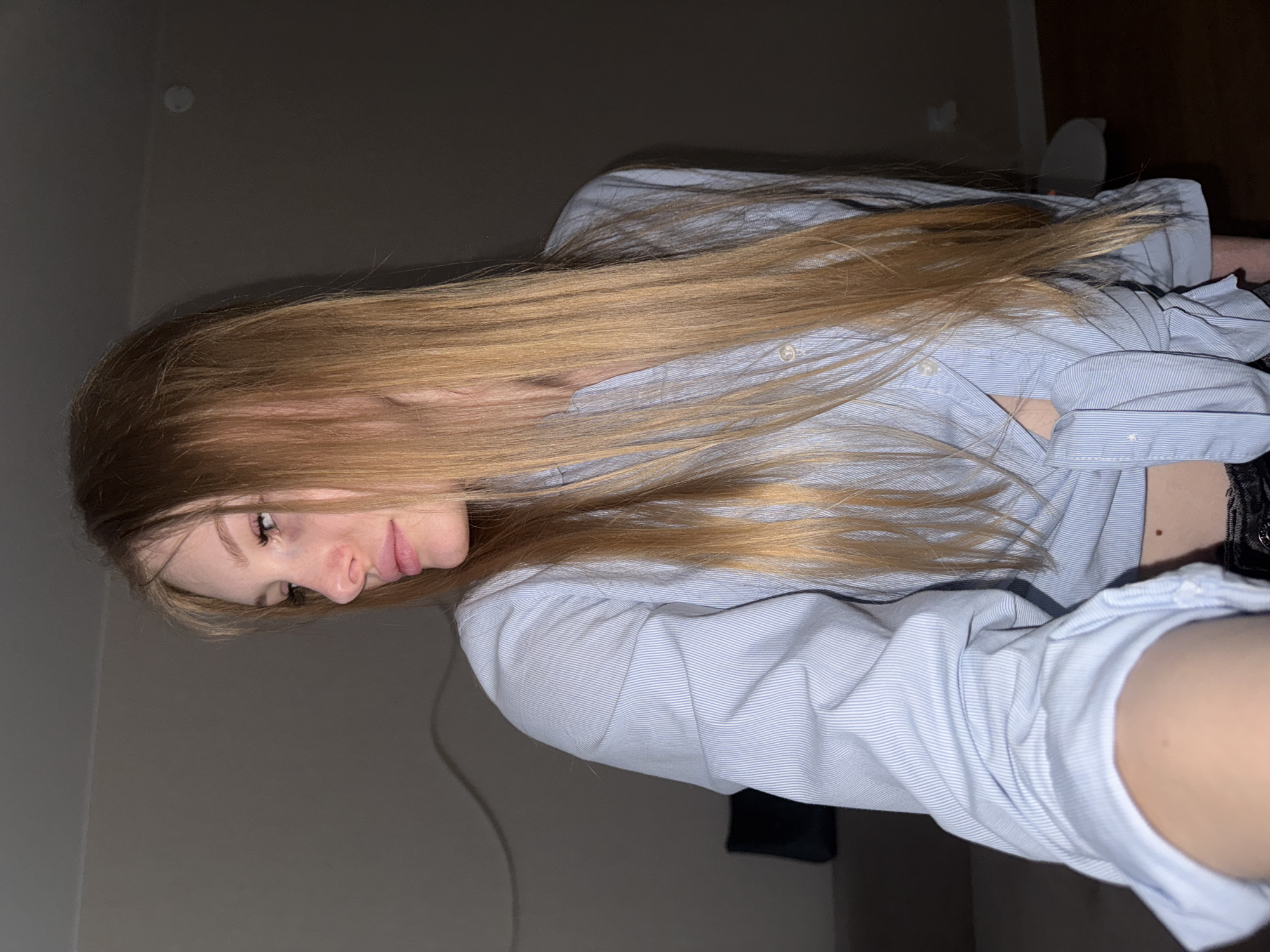 A young woman with long, blonde hair is shown from the shoulders up, looking to her left. She is wearing a light blue, pinstripe button-down shirt, partially unbuttoned, with the sides tied at her waist. Her skin is fair, and she has noticeable pinkness on her cheeks. The background appears to be an indoor residential setting, dimly lit, with a hint of a couch and wall in the muted background. There are no identifiable landmarks or elements that suggest a specific location within the city of Perm, Russia.