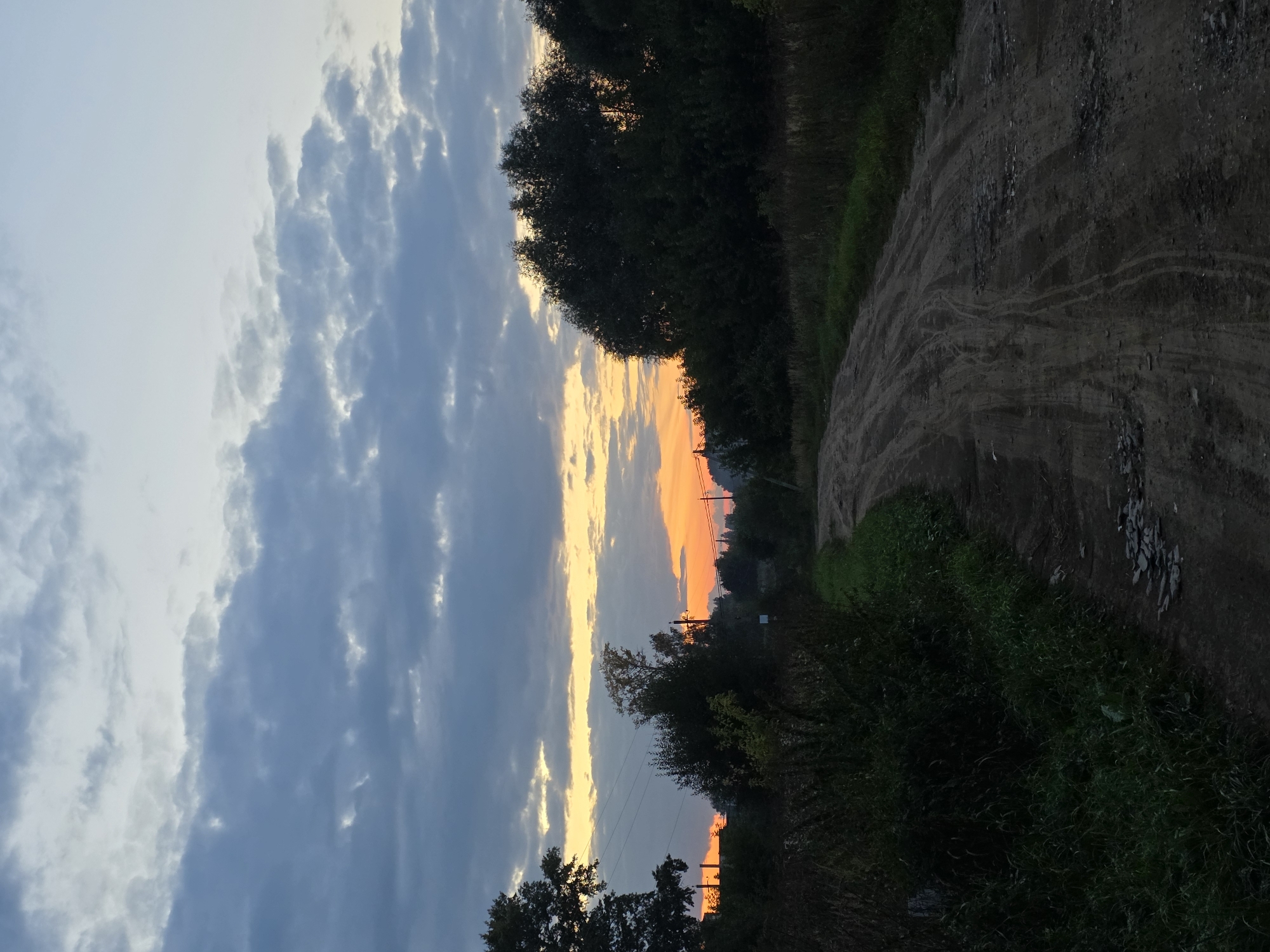 A dirt road curves through a rural landscape under a cloudy sky at dusk. The sky exhibits a gradient of colors from pale blue at the top to orange and yellow near the horizon, where the sun is setting. Dark silhouettes of trees line the sides of the road, with some visible structures and power lines in the distance on the left. The road surface is rough, with visible tire tracks and scattered stones. Tall grasses and vegetation grow along the edges of the road. The overall impression is that of a quiet, unpaved road in a village setting during twilight.