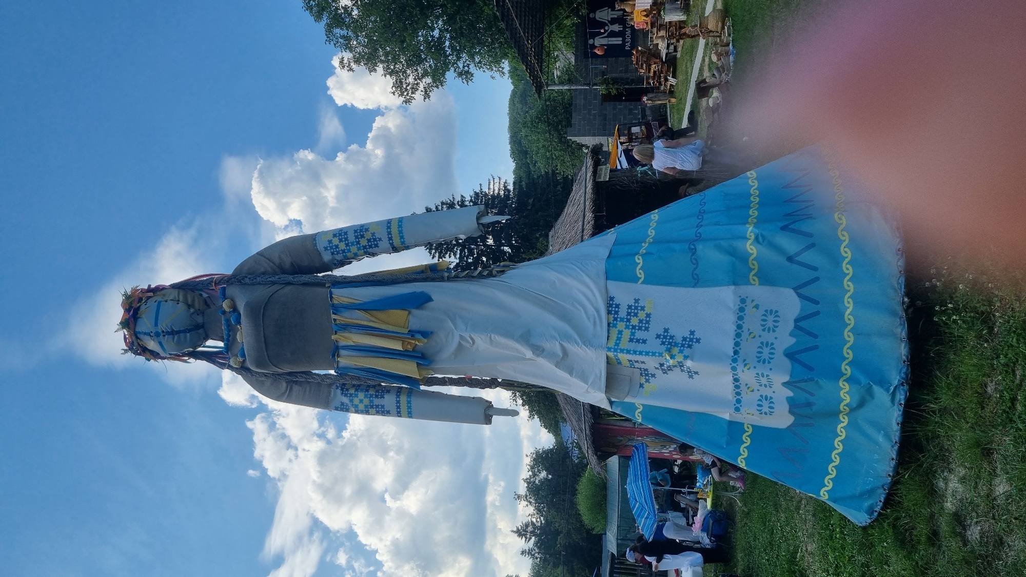 The image displays a colossal, anthropomorphic doll, towering against a bright blue sky with scattered white cumulus clouds, in an outdoor setting identified as Melnyky, Ukraine. The doll is fashioned in the likeness of a woman, with a grey upper torso and long white sleeves adorned with blue and yellow embroidered patterns. Its head is covered with blue fabric, featuring a cross-like design, and it wears a wreath of leaves and flowers, along with long braided hair and blue beaded necklaces. Yellow and blue fabric strips hang from its waist. The lower body of the doll forms a wide, bell-shaped skirt: an upper white section features a large blue and yellow cross-stitch pattern, while the lower section is blue with a yellow wavy line and a blue zigzag pattern.
In the mid-ground to the left, several individuals are gathered around a table shaded by a blue and white striped umbrella. To the right, a young child in a white outfit walks away from the camera, moving toward an area with market stalls. These stalls display various goods, including wooden crafts and pottery, and one sign features Ukrainian text "РАЗОМ СИЛУ" (Together strength) accompanied by pictograms representing a family and a hand holding a heart. The background includes numerous trees and simple structures, some with thatched roofs, suggesting a market or festival environment. The ground beneath the doll is covered with grass and patches of dirt. A reddish, out-of-focus blur, likely a finger, is visible in the lower right corner of the frame.
