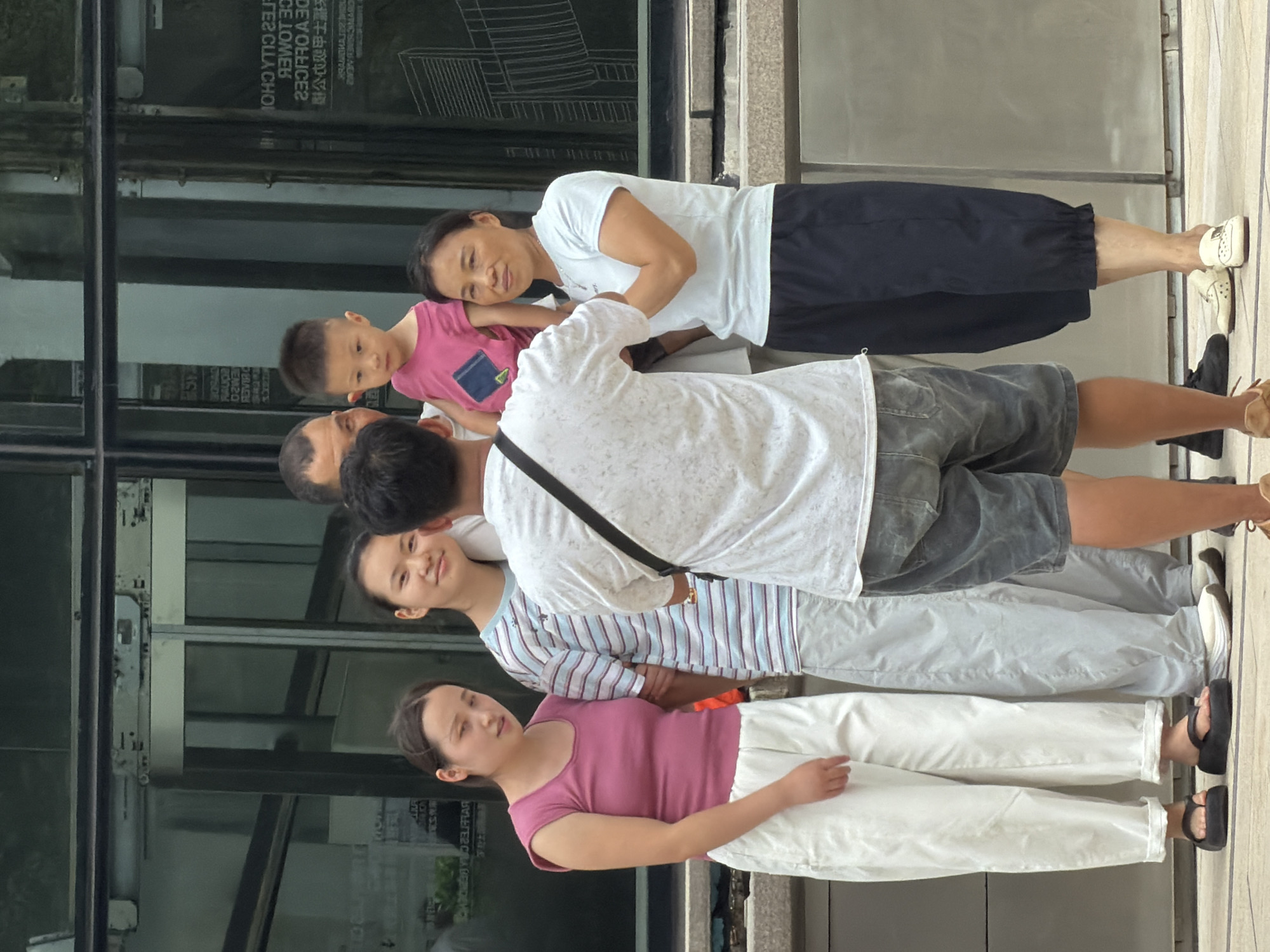 A group of six individuals, appearing to be of Asian descent, are posing for a photograph outdoors in Yuzhong District, China. The setting is in front of a modern building with large glass windows. The group consists of three adults and three children.
On the left, a young woman with dark hair tied back is wearing a sleeveless pink top and loose white pants. She is barefooted and looking towards the camera. Next to her stands a teenage girl with dark hair, wearing a blue and white striped t-shirt and light gray pants. She is smiling at the camera.
In the center, a man with short dark hair is positioned facing away from the camera, appearing to take the photograph. He is wearing a white t-shirt and grey shorts. A young boy in a bright pink t-shirt with a dark square patch is being held by an adult male whose face is partially obscured. The boy is looking towards the camera.
To the right of the group, a woman with dark hair pulled back is wearing a light blue t-shirt and dark blue cropped pants. She is looking towards the camera with a neutral expression. She is wearing white slip-on shoes. Another man, likely the one holding the child, has short dark hair and is wearing a light colored t-shirt.
The ground appears to be paved with concrete tiles. The building behind them has a reflective glass facade with some architectural drawings or designs visible on the interior glass. The lighting suggests it is daytime.