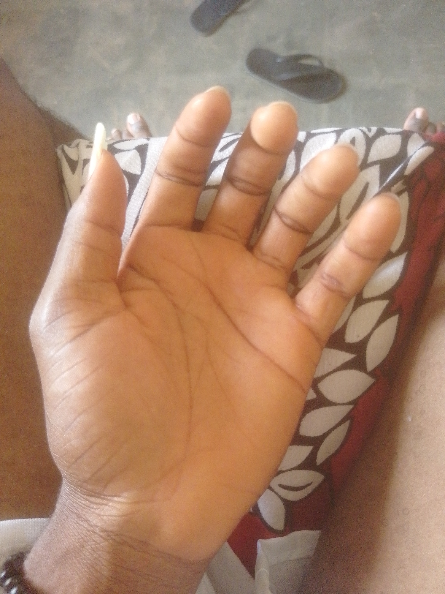 In Majemu, Nigeria, an individual's left hand is prominently displayed, palm facing upwards, against a patterned fabric. The skin tone is dark, and the hand exhibits visible lines and creases. One fingernail, on the index finger, appears unusually long and light-colored. Several fingers are extended, with the thumb slightly bent inwards. In the background, a dark-colored flip-flop is visible on a concrete-like surface, suggesting an outdoor or semi-outdoor setting. The fabric beneath the hand features a repeating pattern of white, stylized leaf shapes on a dark background, with a maroon border.