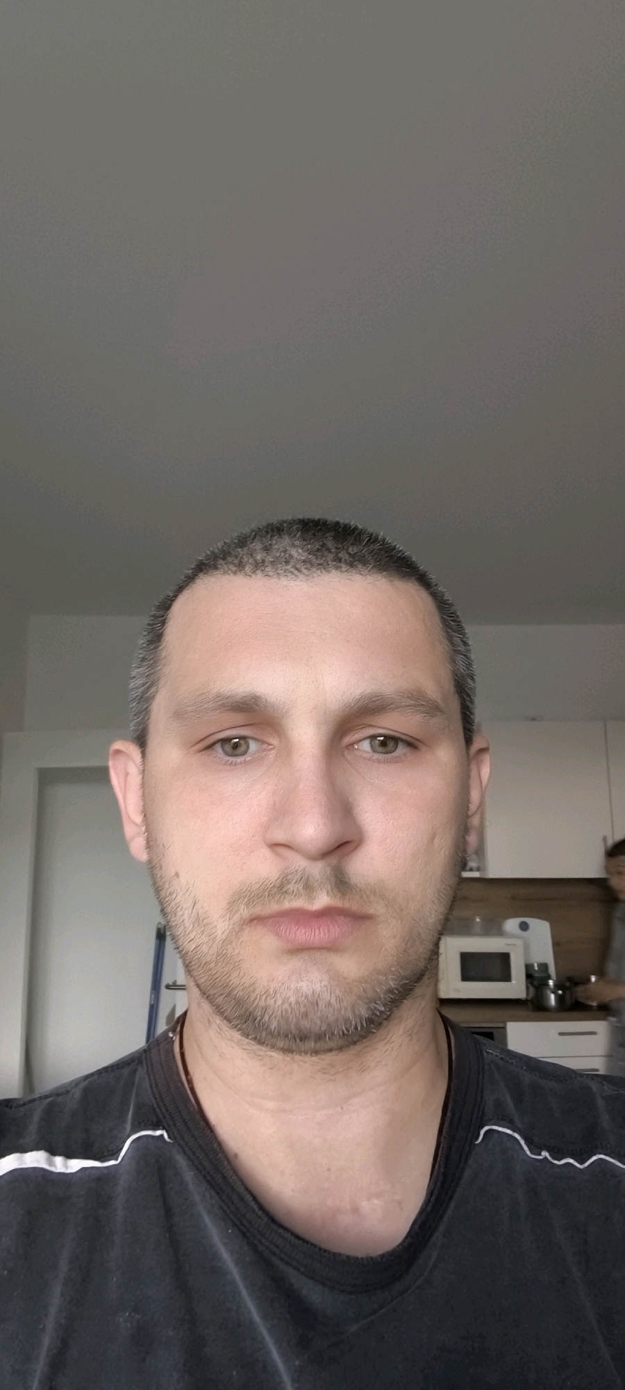 This is a head and shoulders, medium close-up, portrait-style photograph of a man in his late 20s or early 30s with short, dark hair, hazel eyes, and light stubble. He is wearing a dark t-shirt with a white stripe detail on the collar. The background is a blurred interior scene with a white wall, cabinetry, a microwave, and a person in the distance. The lighting is neutral and appears to be natural light from a window or overhead source. The image was taken indoors, possibly in a kitchen or living area. There are no discernible landmarks to indicate the specific location within Wolgast, Germany.