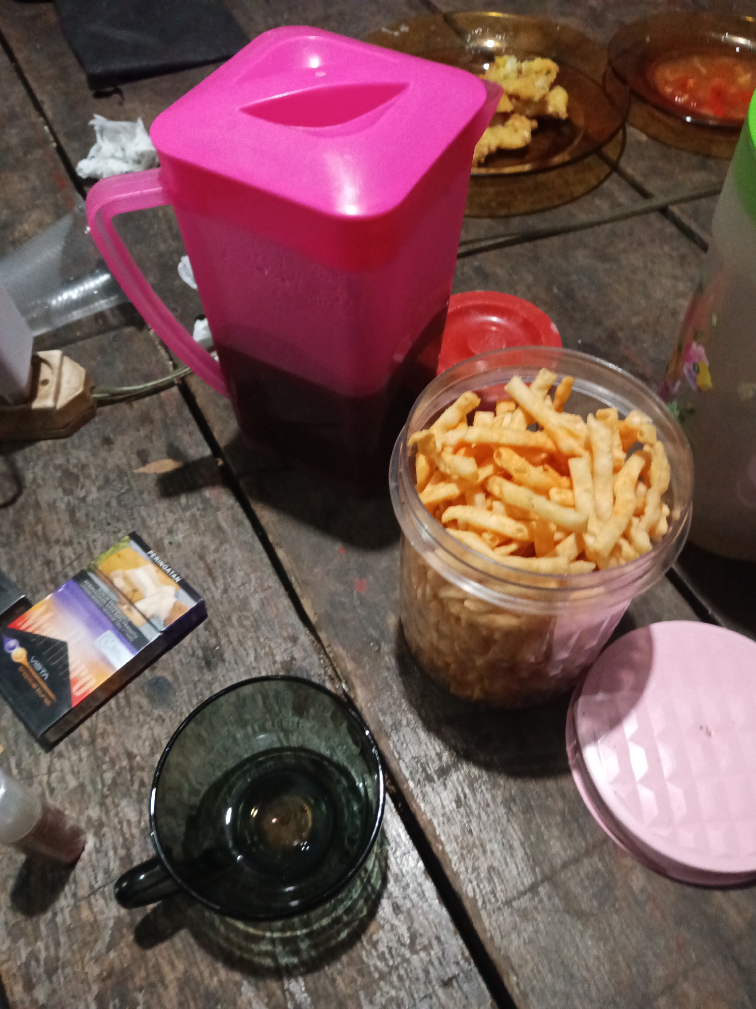 The image depicts a dimly lit, rustic wooden table with various objects arranged upon it. In the center of the frame, a bright pink pitcher holds a dark liquid, likely a beverage. To its right, a clear plastic container is filled with small, golden-brown snack sticks. Adjacent to the snack container is a pink lid. In the foreground, a dark, tinted glass mug sits to the left of the frame. A pack of cigarettes, labeled "VISTA," is placed near the mug. A small bottle with a white cap is visible to the left of the mug, partially obscured. In the background, there are two brown plates, one containing fried food items and the other a reddish sauce or dip. The overall setting suggests a casual gathering or a moment of relaxation, potentially in a home or outdoor setting in Tumampua, Indonesia, given the context provided. The lighting is dim, with shadows cast by the objects, indicating an indoor or evening scene.
