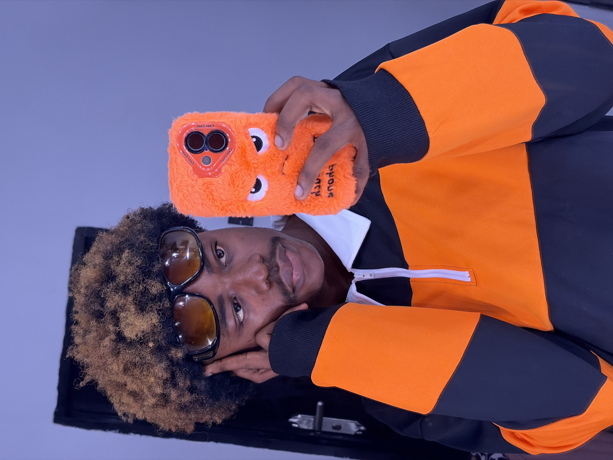 A man is shown from the chest up holding an orange furry phone case with cartoon eyes and an orange ring around a "camera". He is wearing a black and orange pullover sweater with a white collar and zipper. He has brown tinted sunglasses, brown and black hair and is possibly in a city in Nigeria. The image is rotated at an angle making it difficult to determine the environment.