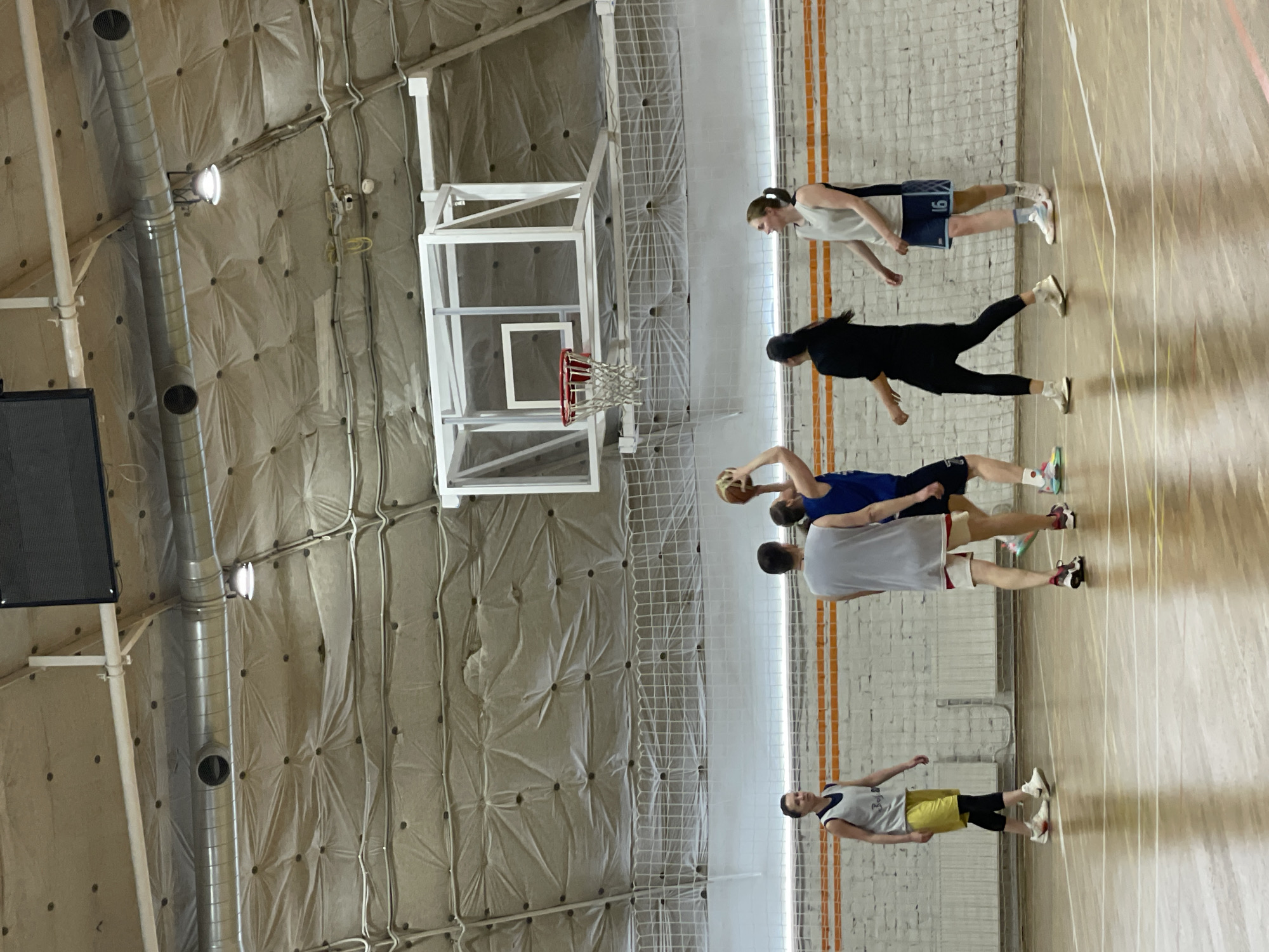 The image captures a basketball court inside a gymnasium. Five people are visible, seemingly engaged in a practice or game. One player in blue is holding a basketball, while others are in various positions around them. The court is marked with painted lines on a wooden floor. The gymnasium features a basketball hoop and a wall with a textured surface. Based on context clues (orange vertical lines in the background) combined with architectural features, it is probable this picture was taken at the Trudovye Rezervy Sports Complex on Ulitsa Glinki in Saint Petersburg, Russia.
