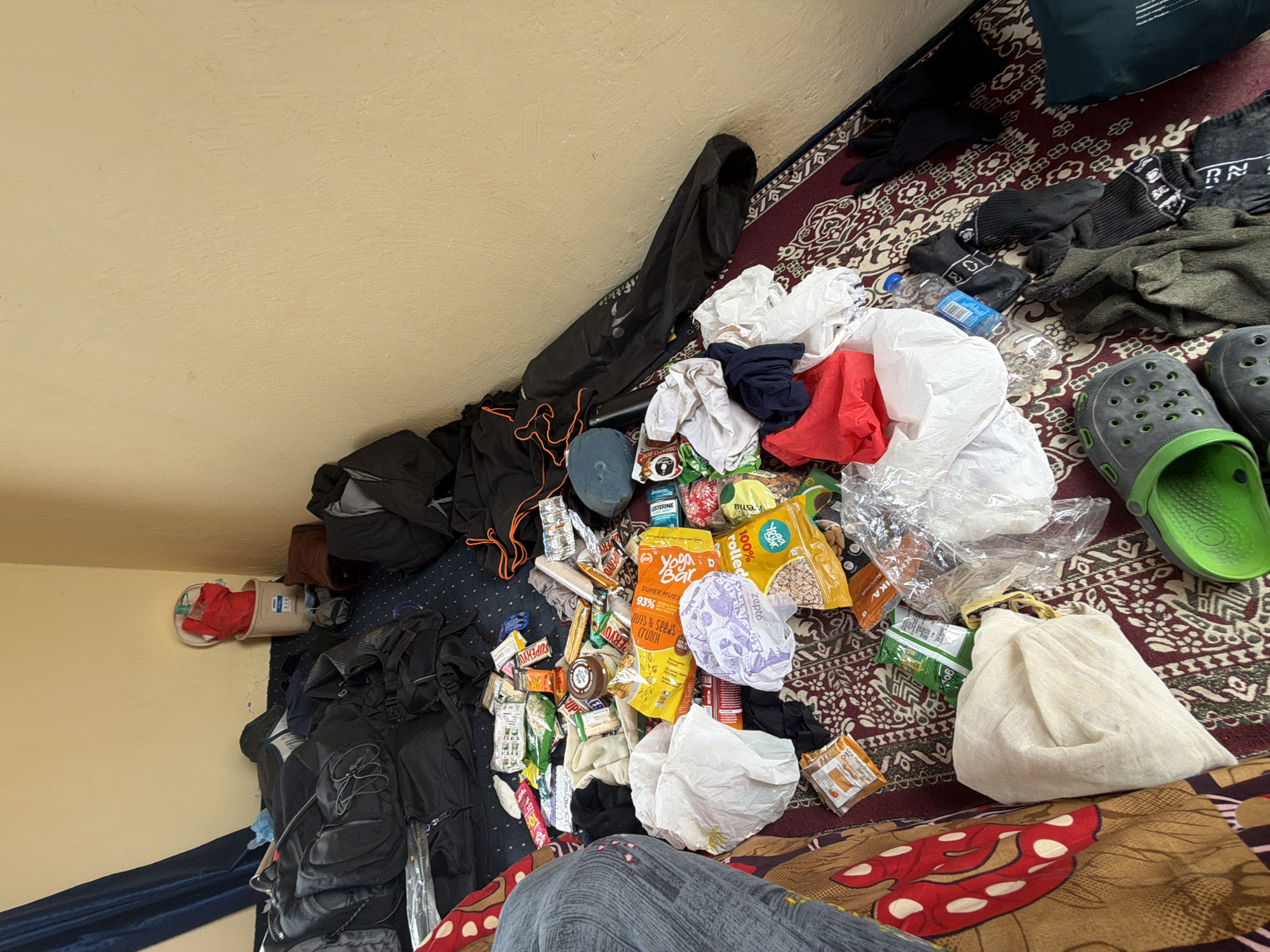 The image depicts a cluttered room with a collection of backpacks, clothing, food items, and personal belongings scattered across a patterned rug. The walls are painted a light, neutral color, and a dark blue curtain hangs on the left side of the frame. In the background, a white bucket with a red item inside sits near the wall. The floor is covered with a red and white patterned rug, upon which numerous items are spread. These include several large black backpacks, various articles of clothing such as socks and what appears to be a red shirt, and a significant number of food packages, including what look like snack bars, cereal boxes, and bags of nuts. There are also some plastic bags, a water bottle, and a pair of green Croc-style shoes. The scene appears to be a disarrayed personal space, likely a bedroom or living area, where items have been unpacked or are in the process of being organized. The location is identified as Karzok, India.
