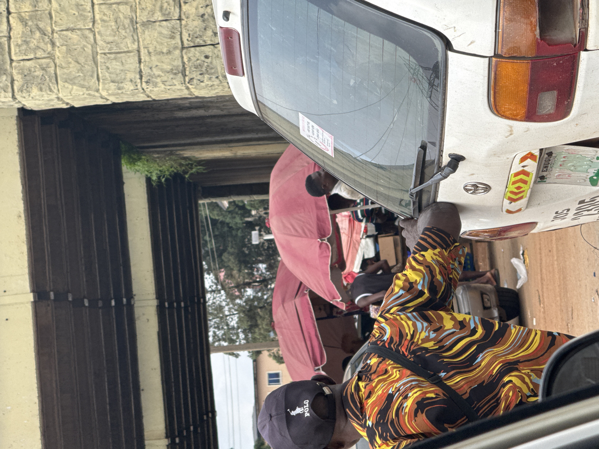 The image captures a bustling street scene under an overpass in Orji, Nigeria. In the foreground, the back of a white Toyota car is visible, with a sticker on its rear window and a license plate indicating "ENUGU" and "GOVERNMENT STATE." A person wearing a colorful, wavy-patterned shirt and a black baseball cap with the "POLO" logo is in the mid-ground, facing away from the camera. Behind them, two red umbrellas are set up, likely for market stalls, with people interacting and standing around. The overpass structure is prominent, made of concrete and steel beams, with brickwork on one side. The environment suggests a typical urban setting with ongoing street activity and possibly a local market.