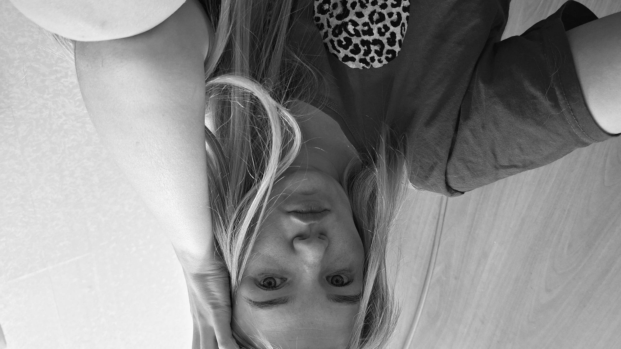 A black and white, medium shot, selfie-style photograph shows a young Caucasian woman from the chest up, looking directly at the camera. She has blonde hair, which falls around her shoulders and partially obscures her face. Her right hand is raised to her head, resting on her forehead. She is wearing a dark-colored t-shirt with a leopard-print design on the left side of her chest. The background consists of a textured wall on the right and a wood-paneled surface on the left. The lighting is direct and even. No specific location in Bolshoy Bukor, Russia, can be identified from the visual cues.