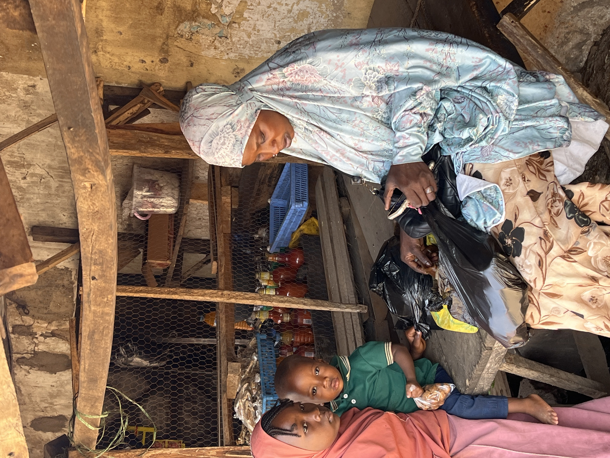 A medium shot of a mother wearing a head covering, a child wearing green, and another child lying together near what seems to be a chicken wire enclosure. The mother is wearing a blue head covering with a floral print. She appears to be in her late 30s or early 40s. She is holding what seems to be a black plastic bag in her left hand and a cell phone in her right. She seems to be looking down at the children. The child lying closest to the camera is wearing a peach-colored head covering and is looking at the camera. The child lying on top of her is wearing a green collared shirt and blue pants, also looking at the camera. The environment is cluttered, with shelves stocked with items, suggesting a shop or storage area. The structural elements such as wooden beams and concrete walls suggest an informal setting. Based on the appearance of the individuals and the environment, it is likely the photograph was taken in Jos, Nigeria, where such scenes are common.
