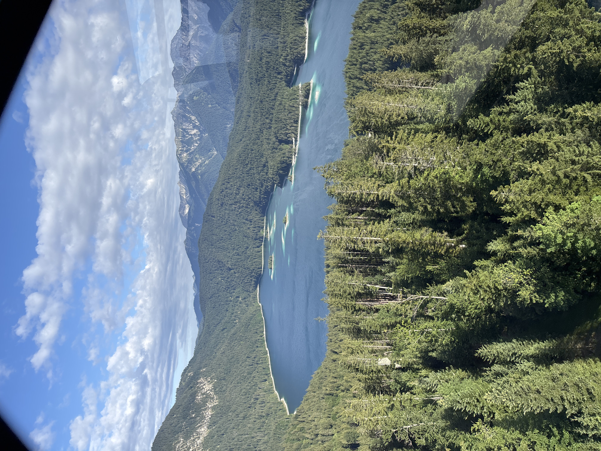 An aerial view shows the Eibsee lake near Grainau, Germany, surrounded by dense coniferous forests and the Bavarian Alps. The lake is a deep blue with lighter turquoise patches indicating shallower areas. Several small, tree-covered islands dot the water. The foreground is dominated by a thick forest of tall evergreen trees. In the background, the rugged, snow-dusted peaks of the Alps rise against a partly cloudy blue sky. The scene depicts a natural landscape on a sunny day.