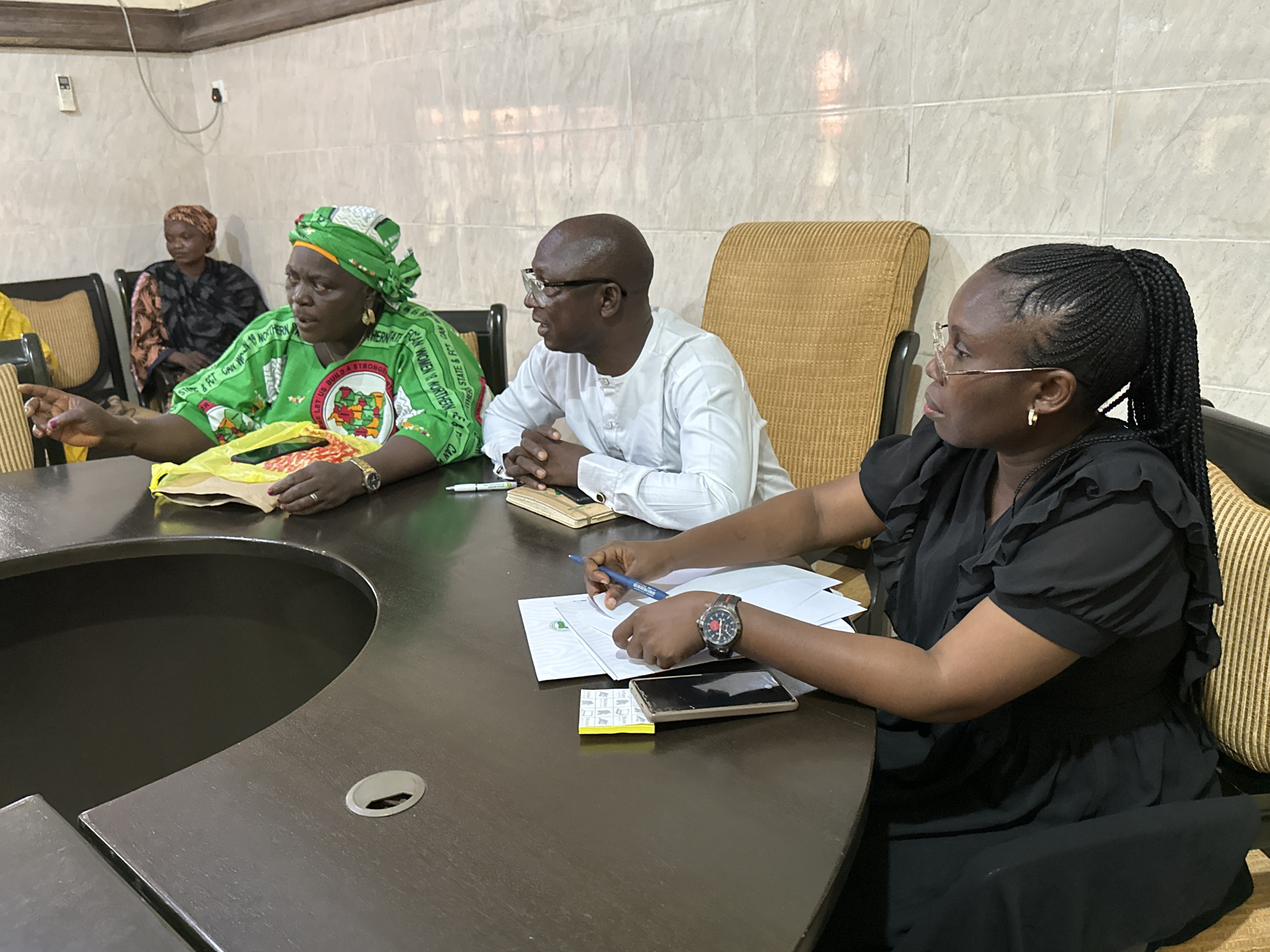 A group of four people are seated around a large, dark brown, oval-shaped conference table. The room has light-colored tiled walls and a wooden ceiling.
In the foreground, a Black woman wearing a black dress with ruffled sleeves and a dark braided ponytail is seated on the right. She is wearing glasses and a gold hoop earring. Her left arm is resting on the table, and she is holding a blue pen, writing on a stack of papers. A smartwatch with a red accent is visible on her wrist. A smartphone is also on the table in front of her.
To her left, a Black man with glasses and a shaved head is seated. He is wearing a white, long-sleeved button-up shirt. His hands are clasped on the table.
To his left, a Black woman wearing a vibrant green headwrap and a green garment with text and a circular emblem is seated. She is gesturing with her left hand towards papers on the table. A yellow plastic bag is also on the table in front of her. She is wearing gold earrings.
In the background, a Black woman in a dark patterned garment and a brown headwrap is seated on the left side of the frame.
The table has a circular cutout in the center. Papers, pens, and a smartphone are scattered across the table. The overall atmosphere appears to be a meeting or discussion. The location is identified as Kabala, Nigeria.