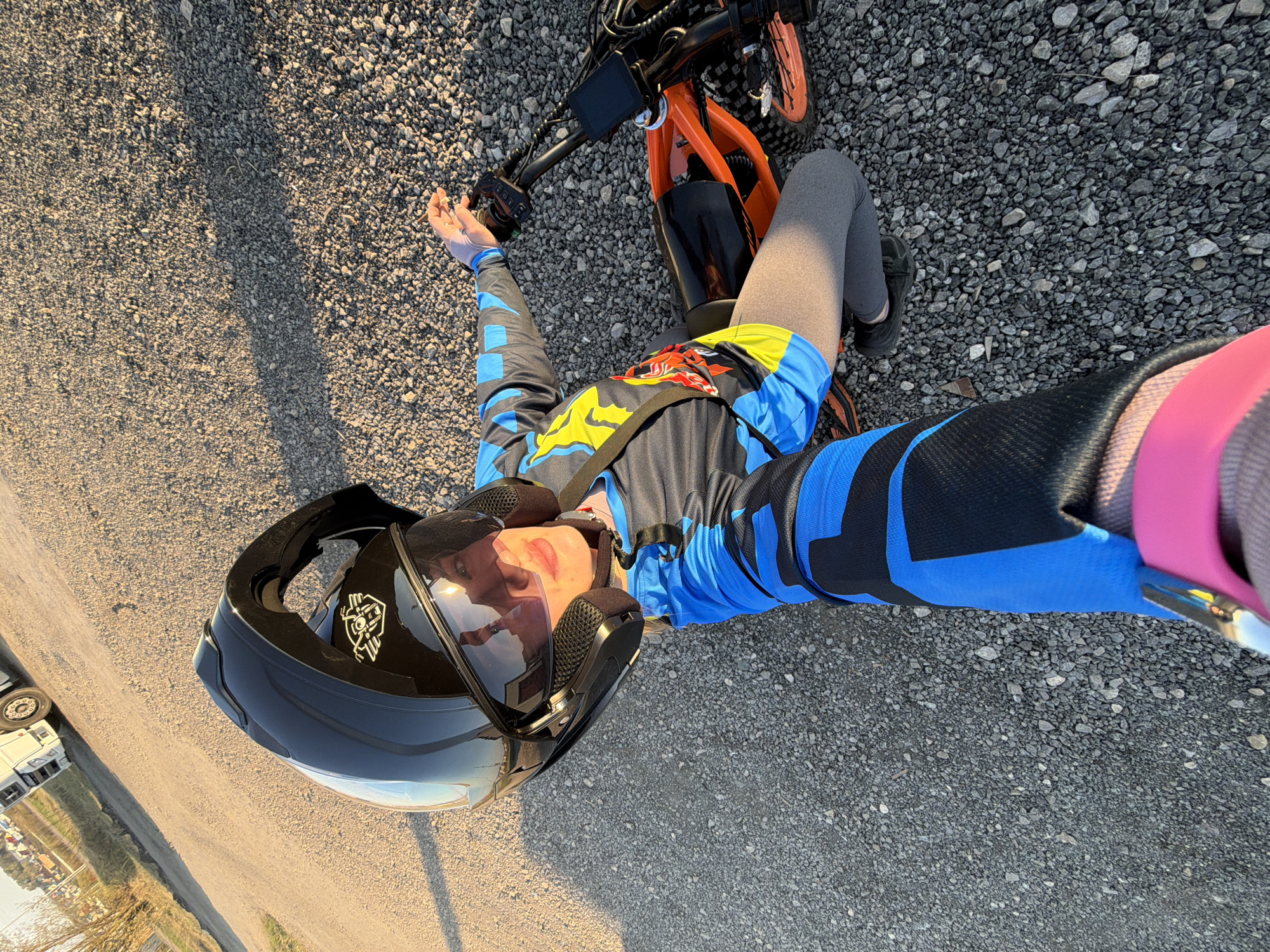 The image shows a person lying on a gravel road, wearing a black motorcycle helmet, a blue and black long-sleeved shirt, gray leggings, and black shoes. The person is next to an orange electric bike. The photo appears to be a selfie taken with the person's arm extended. The background shows a road leading to what seems to be a truck and some buildings. The location is indicated as Азьмушкино, Russia.