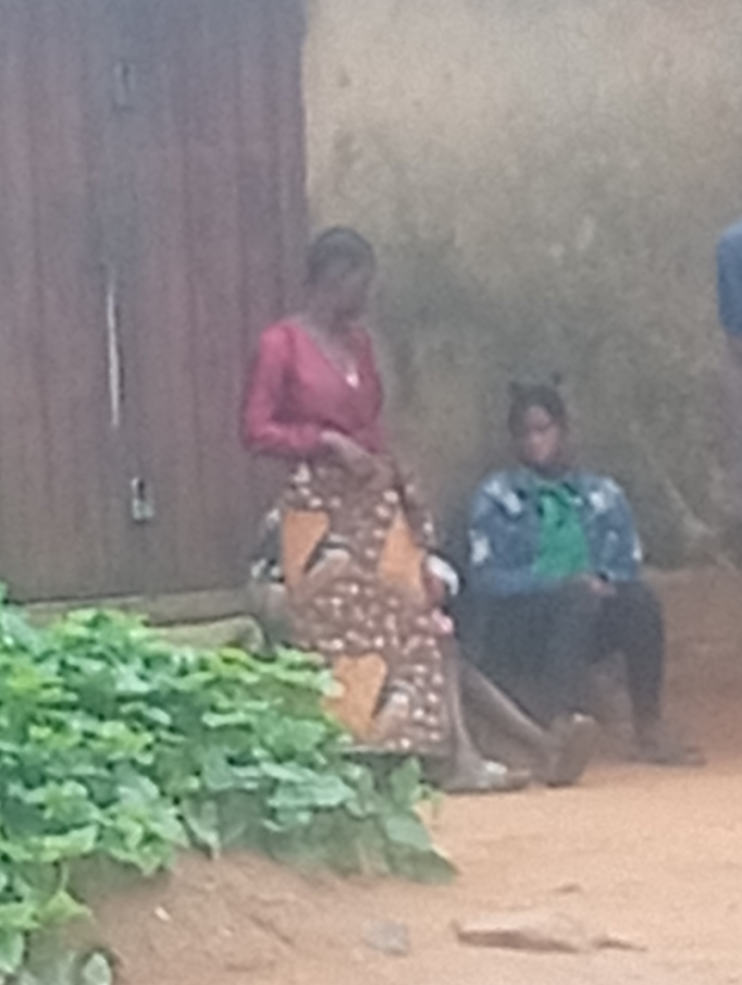 The image shows two women sitting on the ground near a building in a location described only as "Unknown, Unknown". One woman is standing, wearing a red top and a patterned skirt. The other woman is sitting, wearing a blue jacket with a green design over a black top and black pants. In front of them is a patch of green vegetation. Behind them is a wooden door and a stucco wall. The ground appears to be dirt or clay. The overall impression is of a rural or impoverished setting.