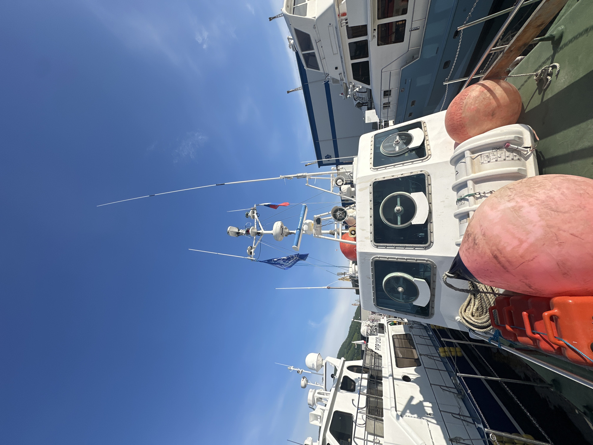 A clear, blue sky looms over a harbor in Petropavlovsk-Kamchatsky, Russia. Several white boats are docked side-by-side. The foreground is dominated by the deck of a boat, featuring two large, pink buoys, several orange canisters, and a coiled rope. The boat's cabin has three large windows with circular shades. A blue flag with white lettering and a white boat with a radar dish are visible on the boat to the left. A Russian flag and another blue flag with white writing are attached to the mast of the boat in the center. In the background, a large, white building with blue trim stands adjacent to the harbor.