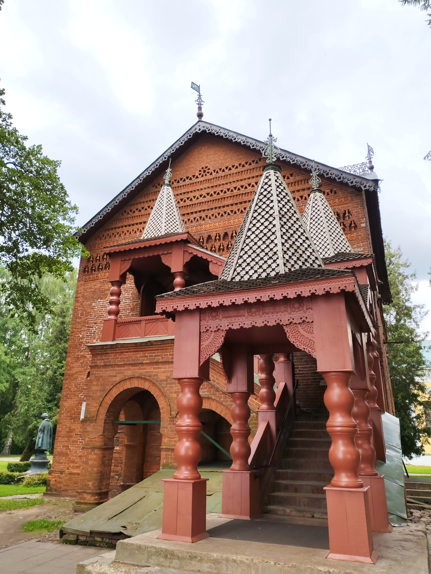 The image depicts a red brick building with a complex wooden roof structure, painted in a dark red hue. The building features arched doorways at its base and a series of decorative wooden porches and balconies. Two conical, patterned roofs rise from the main structure, topped with decorative metal spires. The surrounding environment is green and lush, with trees framing the building. To the left of the building, a dark-colored statue of a man in a coat and hat stands on a pedestal. The sky is overcast with white clouds. The building is likely a historical or cultural site, possibly a church or a museum.