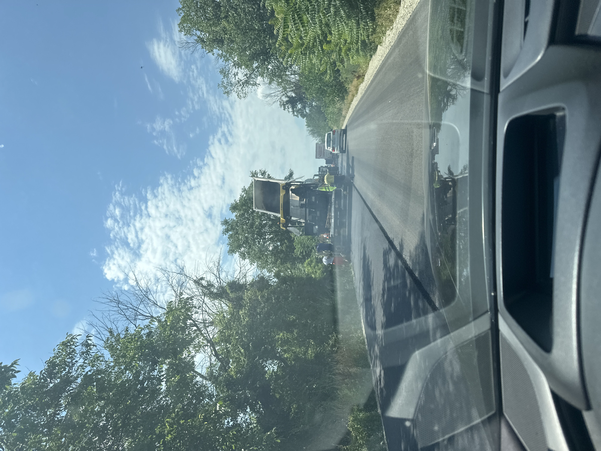 The image, taken from inside a vehicle on a sunny day, depicts road construction activities on a two-lane road bordered by lush green trees. In the foreground, the dashboard of the vehicle is visible, with reflections of the road and trees on the windshield. Further down the road, a large yellow asphalt paver is in operation, laying down new asphalt. Several construction workers in high-visibility vests are present around the paver, some appearing to be directing the material or operating machinery. A white passenger car is visible in the distance, following behind the construction equipment, indicating that traffic is being managed around the work area. The sky is blue with scattered white clouds, and the overall environment suggests an outdoor construction site on a clear day, likely in Polikraishte, Bulgaria, given the context.