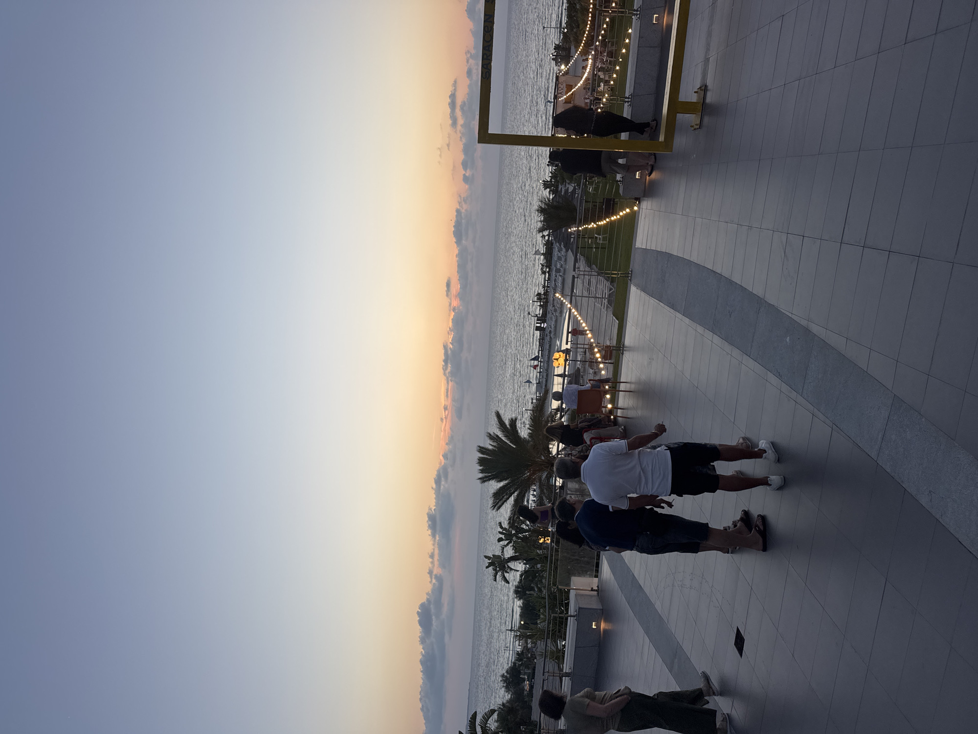 In Isola delle Femmine, Italy, as dusk settles over the Tyrrhenian Sea, a group of people gather on a tiled terrace overlooking the coastline. The sky transitions from a pale blue at the zenith to hues of orange and pink near the horizon, with scattered clouds adding texture. The sea stretches out, calm and reflecting the fading light. Palm trees and other vegetation dot the landscape beyond the terrace, interspersed with what appear to be beach chairs and umbrellas. String lights create a festive ambiance along the edge of the terrace, illuminating pathways and seating areas.
Several individuals are visible on the terrace. In the foreground, a woman in dark pants and a beige top stands with her back to the camera, looking out towards the sea. To her left, two men are engaged in conversation, one in a dark blue shirt and jeans, the other in a white polo shirt and black shorts. Another man in a white polo shirt and dark shorts stands facing away from the camera. Further into the scene, other figures can be seen mingling and seated at tables. On the right side of the frame, a yellow frame, possibly part of a photo booth or decorative element, stands prominently. The tiled floor of the terrace features a repeating geometric pattern. The overall atmosphere suggests a relaxed evening gathering, perhaps at a restaurant or resort, enjoying the seaside scenery at twilight.