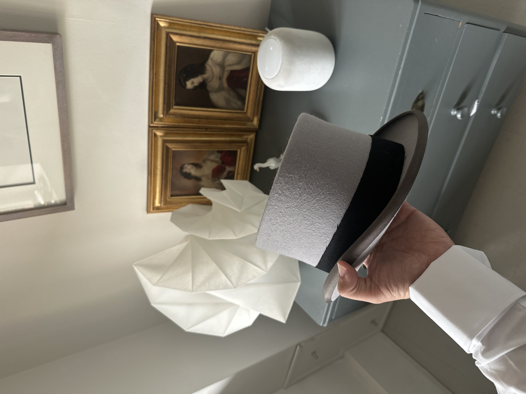 An indoor scene depicts a human hand holding a light gray felt top hat with a black ribbon band. The hand, exhibiting fair skin and wearing a white shirt with a crisp cuff, is positioned in the lower-left. The background features a dark gray dresser with silver drawer pulls, upon which rests a white smart speaker, a small white figurine, and two white geometrically folded paper sculptures or lampshades. Above the dresser, two ornate gold-framed paintings, depicting figures in historical attire, are displayed, with a larger framed object hanging above them on a light-colored wall. The floor and additional wall sections are also light-colored. The specific location within the city of London, United Kingdom, cannot be determined from the visual information provided.