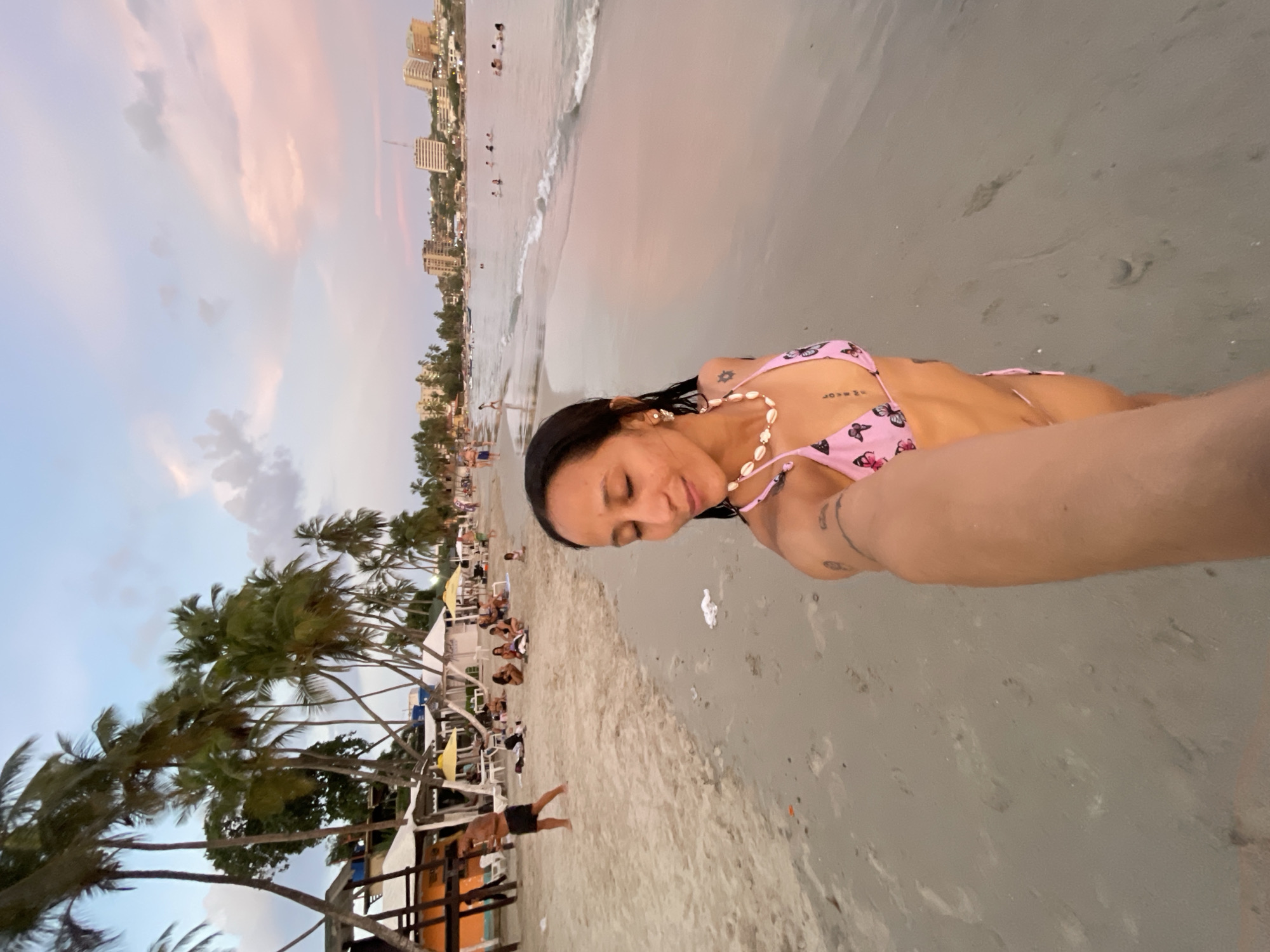 A woman in a pink butterfly-patterned bikini takes a selfie on a beach at sunset in Pampatar, Venezuela. She is wearing a necklace made of seashells and has tattoos on her arm and torso. The beach is sandy with palm trees and small buildings in the background. The water is calm with gentle waves, and people are swimming and relaxing on the shore. The sky is a blend of blue, pink, and orange hues, indicating the sun is setting.
