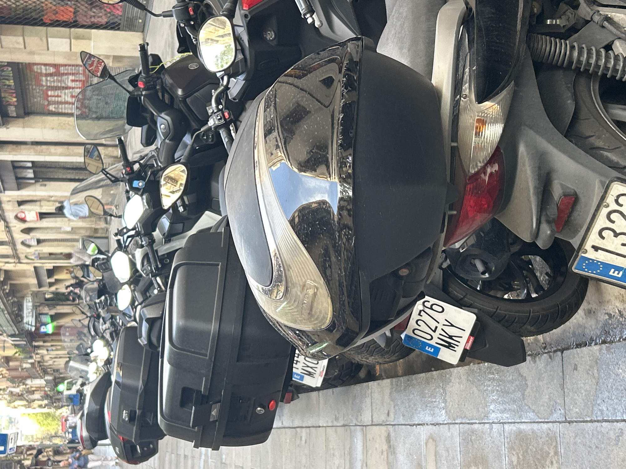The image captures a longitudinal view of multiple scooters parked in a row on a paved street or sidewalk in an urban setting, identified as Barcelona, Spain. The scooters are predominantly dark-colored, with several featuring large, black top cases for storage, clear windshields, and rearview mirrors. The perspective is slightly angled, showing the side and rear of the closest scooters, extending into a blurred line of more parked bikes in the distance.
The ground is paved with light-colored, rectangular tiles or cobblestones. In the background, blurred buildings with multiple stories and some storefronts, including one with a red corrugated metal shutter pulled down, suggest a busy commercial or residential area. Bright reflections on the glossy surfaces of the scooters, particularly on the headlights and body panels, indicate it is daytime and sunny. These reflections also show distorted views of the surrounding architecture and the sky.
No direct human activity is depicted; the scene focuses solely on the parked vehicles, implying routine urban parking.
Visible text includes several European license plates. The most clearly legible plate belongs to the closest scooter and reads "0276 MKY" with a blue strip on the left containing the "E" for Spain. Another partial license plate "MXC" is visible on a scooter further down the line, and another partially visible plate ends in "13" also featuring the "E" for Spain. Faint text "TÜV" can be seen on the headlight of one scooter. Reflections in the background and on the scooters show some blurred text from shop signs, but it is largely unreadable.