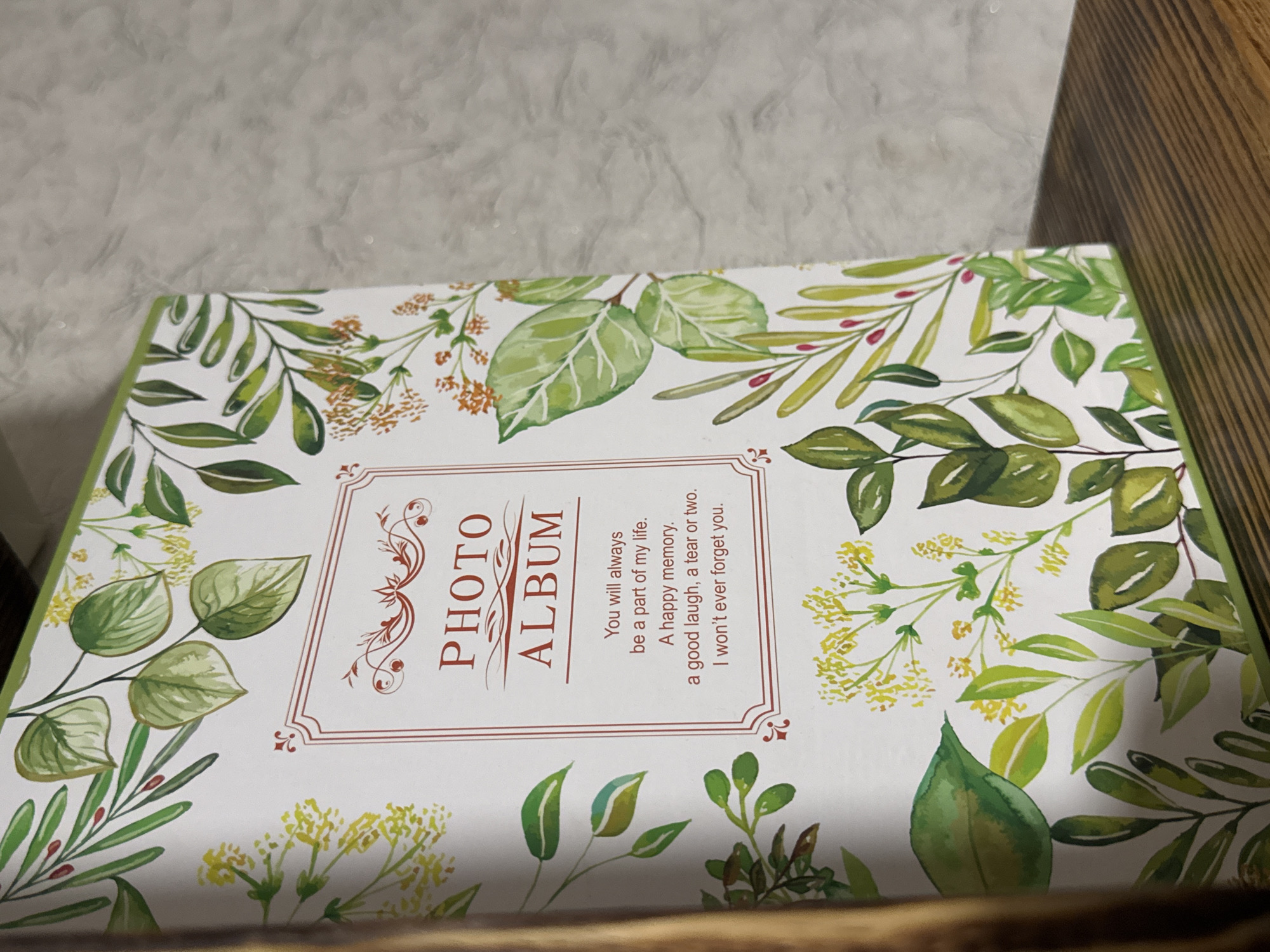 A close-up, eye-level shot shows the cover of a photo album. The album is white and decorated with watercolor illustrations of green leaves and small yellow and orange flowers. A red border frames a central area with the words "PHOTO ALBUM" in red text. Below this, a quote in black text reads, "You will always be a part of my life. A happy memory. a good laugh, a tear or two. I won't ever forget you." The album is positioned on a wooden surface, likely a shelf, with a textured gray background behind it. There are no people or animals visible in the image. The scene is static and appears to be indoors.