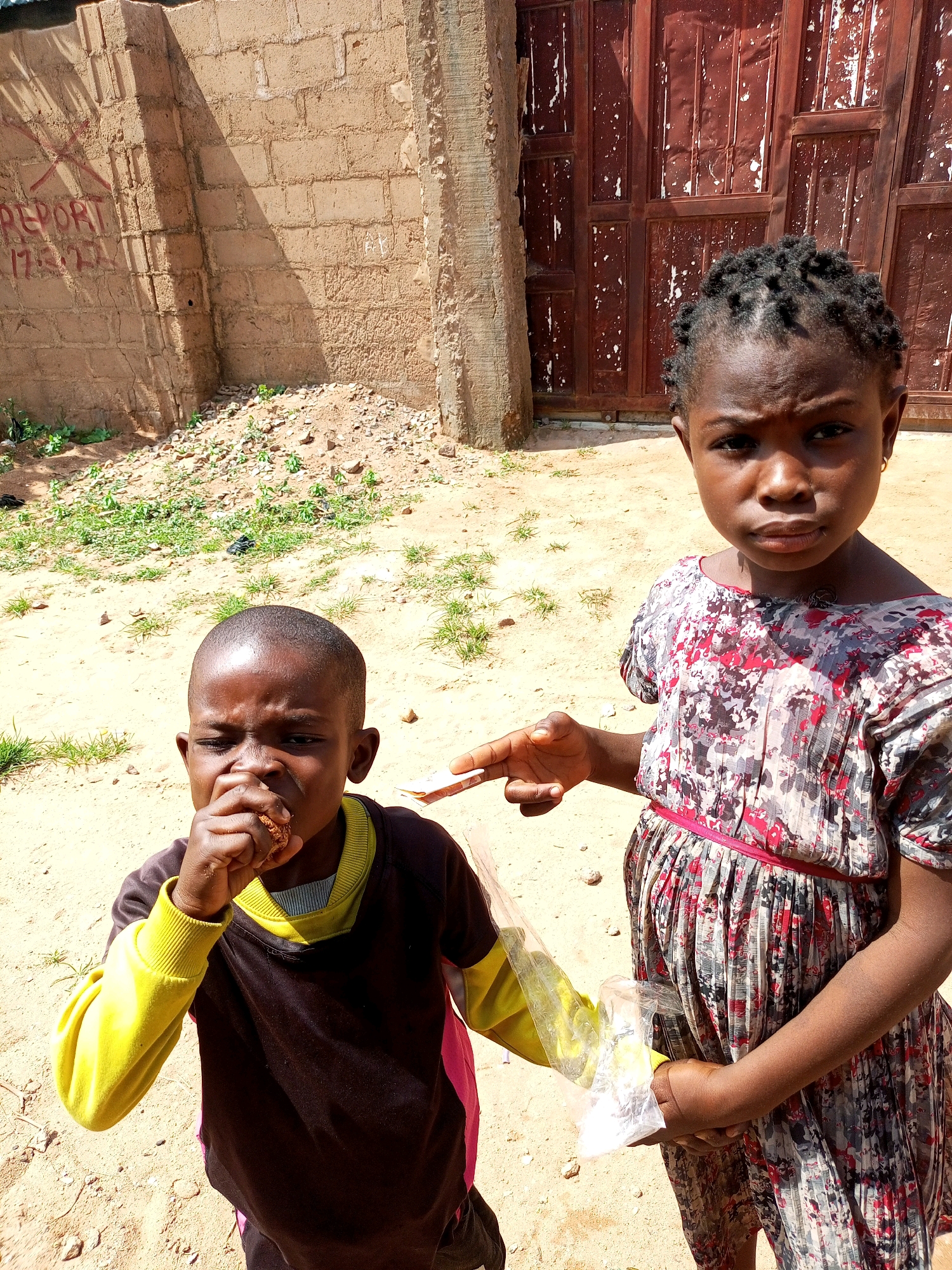 Two young children, a boy and a girl, stand in an outdoor setting in Tempure, Nigeria. The boy, on the left, is eating something he holds in his hand and is wearing a long-sleeved shirt with yellow sleeves and a dark body. The girl, on the right, is wearing a patterned dress and is holding a clear plastic bag with a white substance inside. She is pointing towards the boy with her right hand. Behind them is a brick wall with some graffiti and a large, reddish-brown metal door. The ground is sandy with scattered patches of dry grass and small stones. The lighting suggests it is daytime with bright sunlight casting shadows.