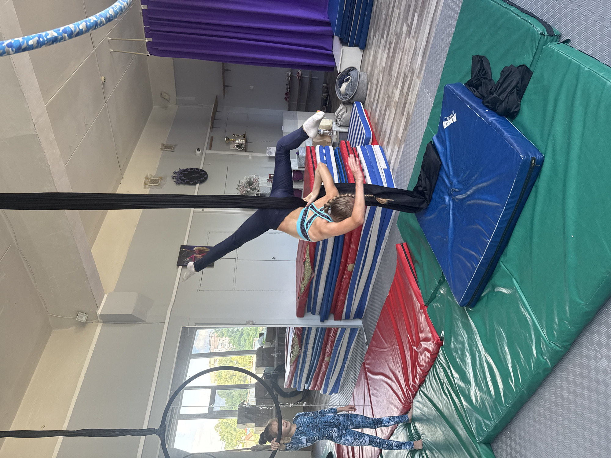 A woman is performing aerial silks in a studio. She is upside down, suspended by black silks, with her legs extended. She is wearing a blue and black sports bra and navy blue leggings. Another person, a child, is on the floor below, holding onto a hoop, and wearing a blue and black patterned jumpsuit. The studio has a purple curtain in the background, shelves with equipment, and a window with a view of trees and buildings. There are also several mats on the floor, including green, blue, red, and blue and white striped mats. The scene appears to be indoors, with artificial lighting. The overall mood seems to be one of athletic activity and practice. There is some text visible on a blue mat that says "DOBLE".
