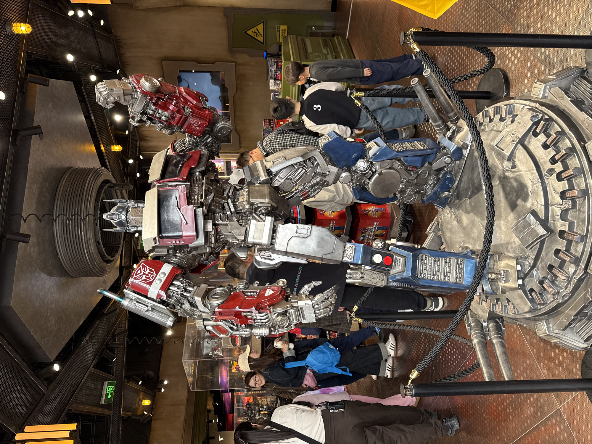 The image depicts a life-size replica of a Transformer character on display, presumably in an indoor attraction or museum. A group of people, including adults and children, are looking at the replica. The Transformer is primarily red, silver, and blue, with a complex, mechanical design. The environment appears to be themed, with industrial-style elements such as metal grates and simulated machinery. A black rope barrier is set up to keep people at a distance from the exhibit. Based on the prompt's instruction, this picture was taken in Tongzhou District, China.
