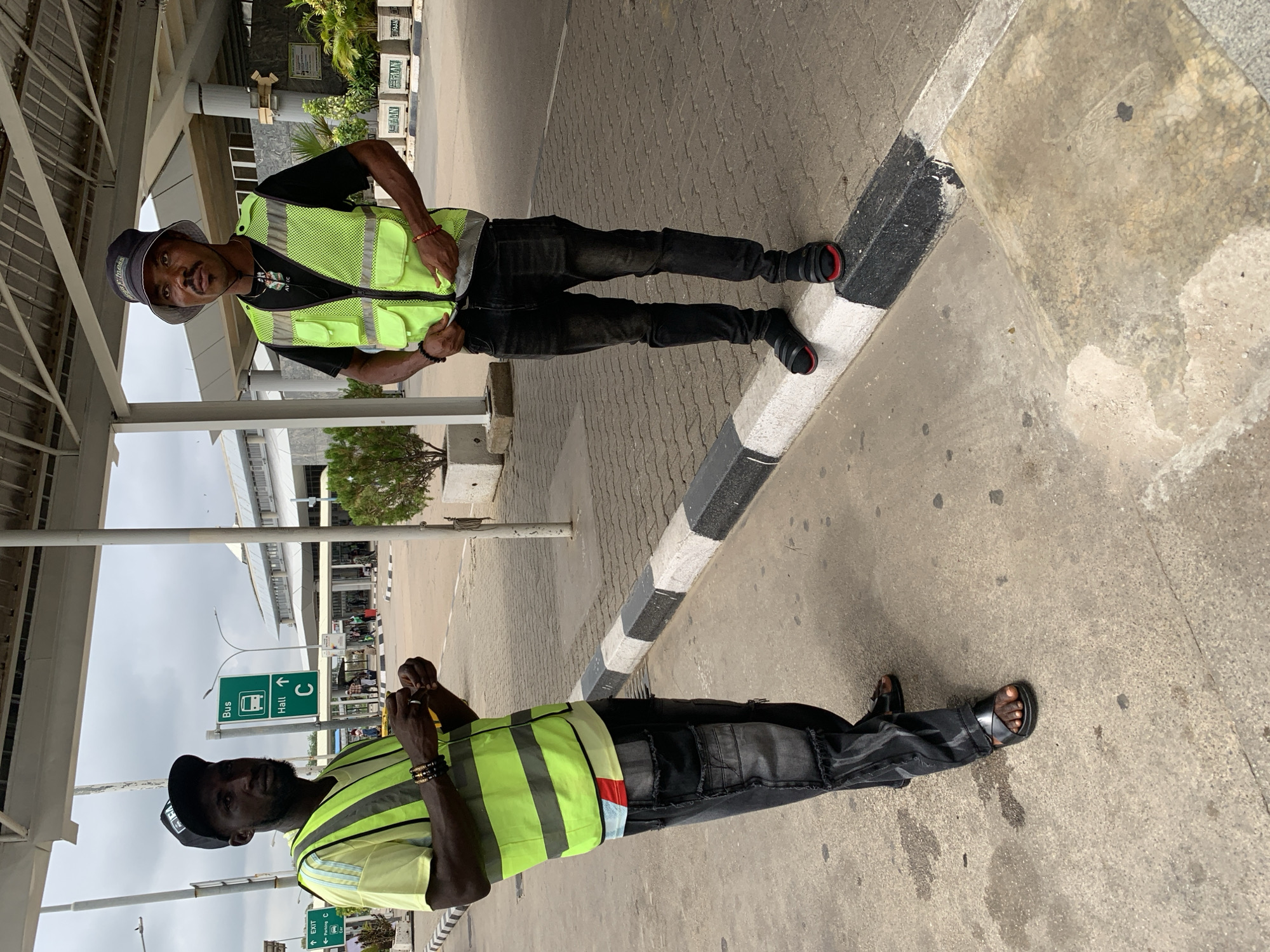 Two men wearing neon yellow safety vests stand on a paved surface next to a road, possibly in Gwaska, Nigeria. The image appears to be taken during the daytime under an overcast sky.
The man on the left wears a dark bucket hat with the word "AVEC" visible, a black t-shirt, dark pants, and black sandals with red accents. He has a light mustache and is looking towards the camera with a slight smile, his hands resting on his vest pockets. He wears red and black bracelets on his left wrist.
The man on the right wears a black baseball cap with "ELI" or "FFL" visible, a black t-shirt, dark pants with textured panels, and black sandals. He has a beard and is looking slightly downwards or forward, holding his hands together in front of him. He wears dark bracelets or rings on his wrists.
They are positioned near a black and white striped curb that separates the paved area from a concrete section. In the background, there is an overhead structure, various buildings, and some greenery. Prominently visible are green traffic signs indicating directions: one reads "Bus" with a bus icon and "Hall" with an upward arrow, and "C" also with an upward arrow. Another smaller sign below it shows "Exit" and "C" with arrows, suggesting a transportation hub like an airport. In the far background, stacked items with the word "HERBAN" are faintly visible.