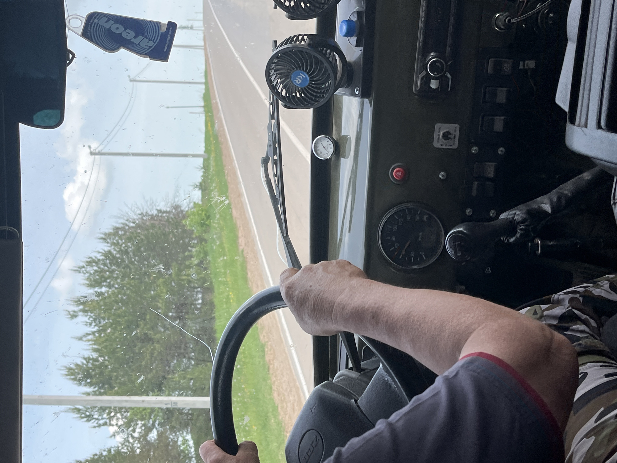 A view from inside a vehicle traveling on a road. The driver's hands are on the steering wheel. The dashboard is visible, with gauges, switches, and two small fans attached. An air freshener hangs from the rearview mirror. Outside the windshield, there is a road with vegetation on both sides. The location appears to be rural, but the city is unknown.