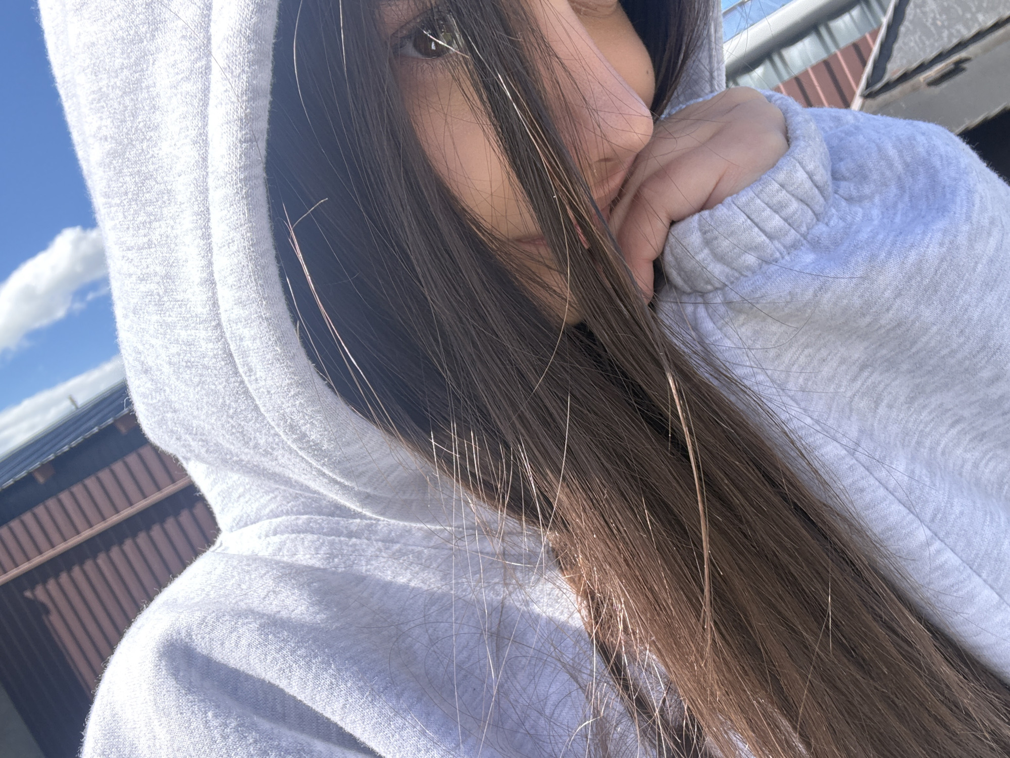 A close-up, angled photograph shows a person wearing a light gray hoodie, with the hood pulled up. The person’s face is partially obscured by long, brown hair. The person's hand is raised to their face. Behind the person is a building with corrugated roofing and brown walls, possibly located in Stepanavan, Armenia, with clear blue sky and a cloud visible. The photograph is taken outdoors during daylight.
