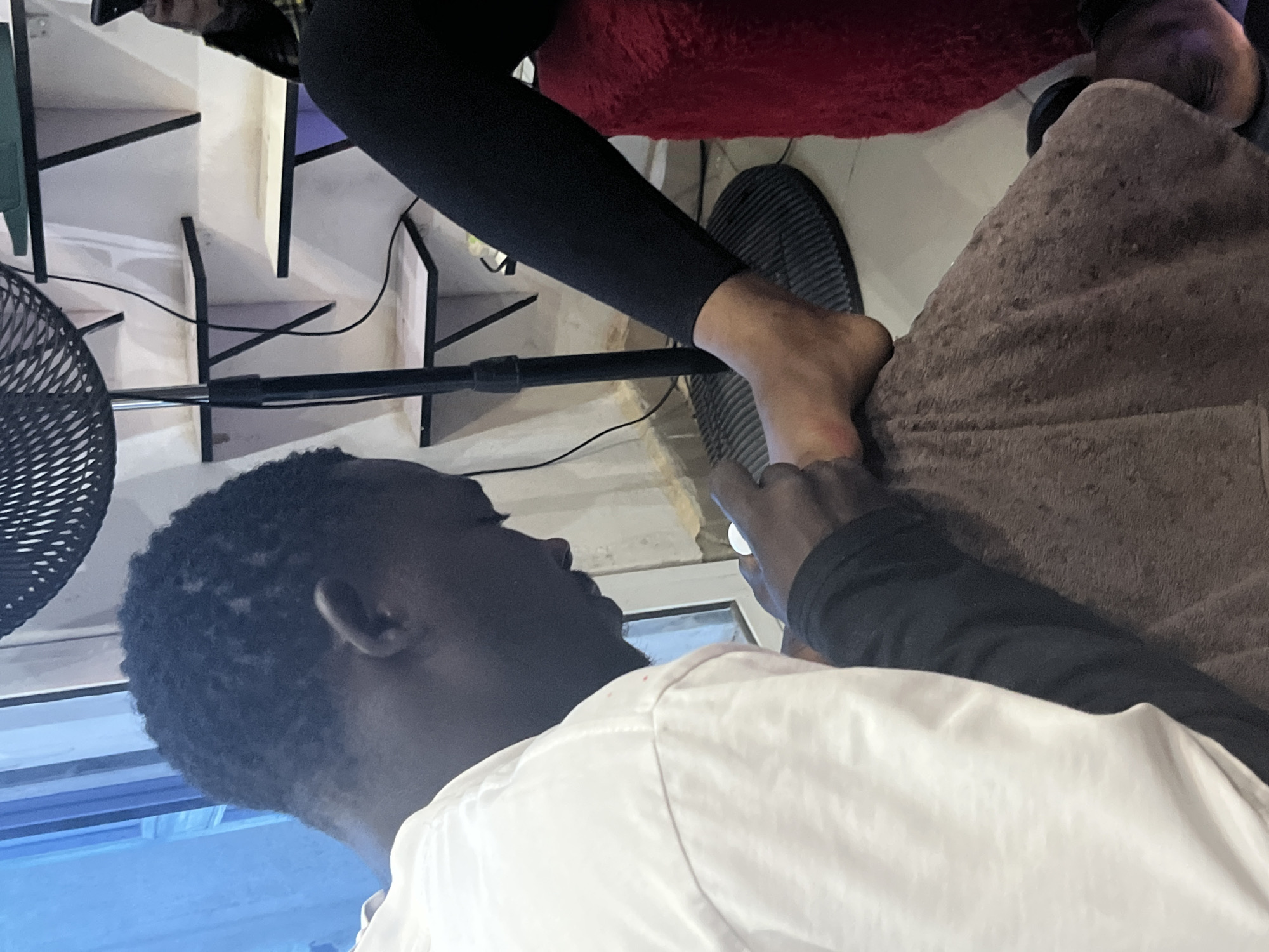 A person, identified as a male with dark skin and short, dark hair, is seated on the ground, wearing a white t-shirt and dark trousers. He is positioned to the left of the frame and is focused on applying nail polish to the toenails of another individual, whose lower legs and feet are visible in the center and right of the frame. This individual is wearing black leggings and is seated on a red, fuzzy stool. A brown towel is placed under the feet being worked on. In the background, a black oscillating fan stands tall. Behind the fan, white shelves are mounted on a wall. The lighting suggests an indoor setting, possibly a salon or a home. The overall scene depicts a person receiving a pedicure. The image is taken in a residential area of Jos, Nigeria.