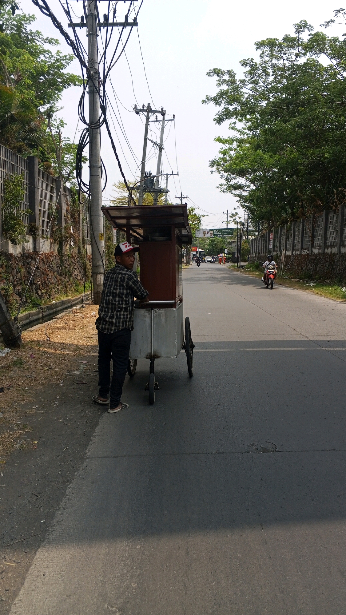 A man in a checkered shirt and a baseball cap stands next to a mobile food cart on a paved road in Semarang, Indonesia. The cart has a silver base with two wheels and a wooden upper structure with a canopy. The man is facing away from the camera, towards the street. The road is bordered by a low wall and vegetation on the left and a concrete wall with a fence on the right. Overhead power lines are visible, supported by utility poles. Several motorcycles are present on the road, with one riding towards the viewer. The sky is overcast.