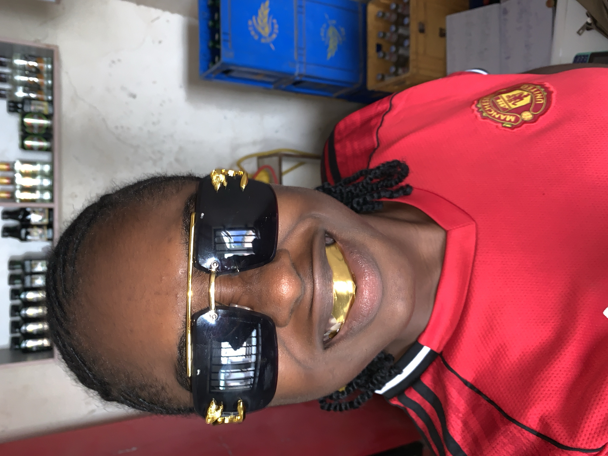 A person with dark skin is prominently featured in a close-up, chest-up portrait, oriented vertically. They are wearing large, stylish black sunglasses with intricate gold detailing on the frames. A gold grill is visible on their upper front teeth, with their mouth slightly ajar. Their dark hair is styled in distinct braids or twists. The individual is wearing a vibrant red Manchester United football club jersey, which features the club's crest and the text "MANCHESTER UNITED" clearly visible on the right side of their chest (viewer's left). Black stripes adorn the shoulder and sleeve area of the jersey.
The background indicates an indoor commercial setting, likely a shop or bar. To the left, shelves stocked with various bottled beverages are visible. In the upper background, several crates filled with bottles can be seen, notably two blue crates displaying a yellow logo resembling a wheat stalk or similar motif. The wall in the background is a light, textured, and somewhat weathered surface. The person appears to be posing for the camera, conveying a sense of confidence and personal style. This scene takes place in New Stadium, Nigeria.