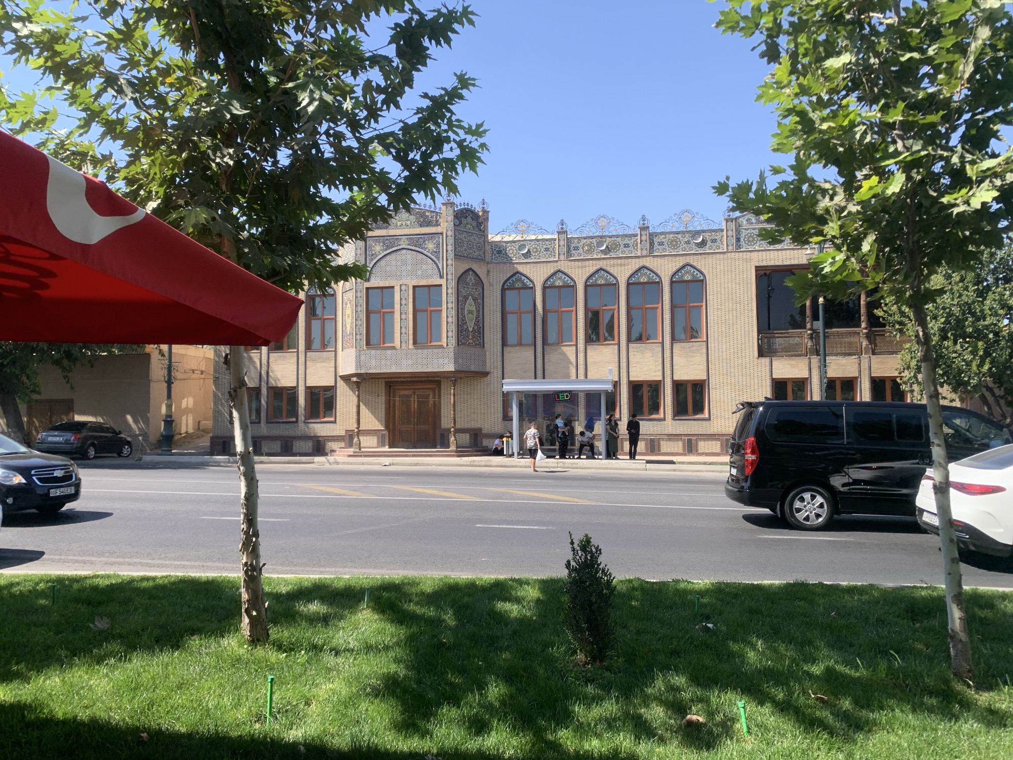 The image depicts a street scene in front of a building with ornate, traditional architecture. The building features a light-colored facade with intricate tilework and arched windows. In the foreground, a red awning is visible, casting a shadow on the street. Several cars, including a black minivan and a white sedan, are parked or driving on the road. A few people are gathered near a bus stop shelter in front of the building, with some individuals sitting and others standing. Trees line the street and a well-maintained grassy area with sprinklers is in the foreground. The sky is clear and blue.