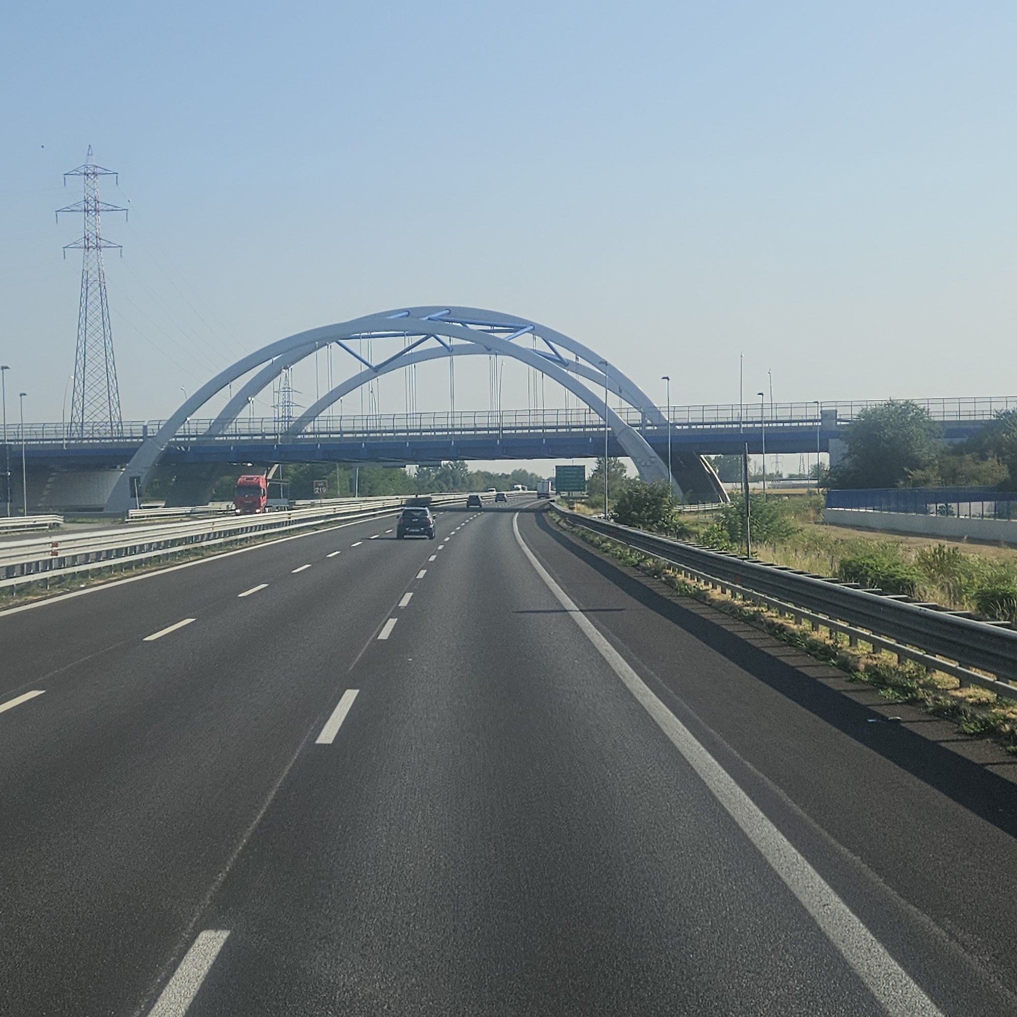 This is a frontal, eye-level photograph taken from a vehicle moving on a highway. The road surface is asphalt with visible white lane markings. A metal guardrail runs along the right side of the road. In the background, a large, modern bridge with twin arched spans, painted in light blue and white, crosses the highway. Below this bridge, a separate, straight blue bridge is visible. To the left of the highway, a tall, lattice-style electrical pylon stands against a clear, pale blue sky. A red truck is positioned on the road beneath the arched bridge, and several cars are also visible on the highway, heading in the same direction as the photographer. The surrounding environment appears to be rural or semi-rural, with sparse vegetation and trees visible beyond the highway. The overall impression is one of a bright, sunny day on a major transportation route. The location is identified as being in the municipality of Montirone, Italy.