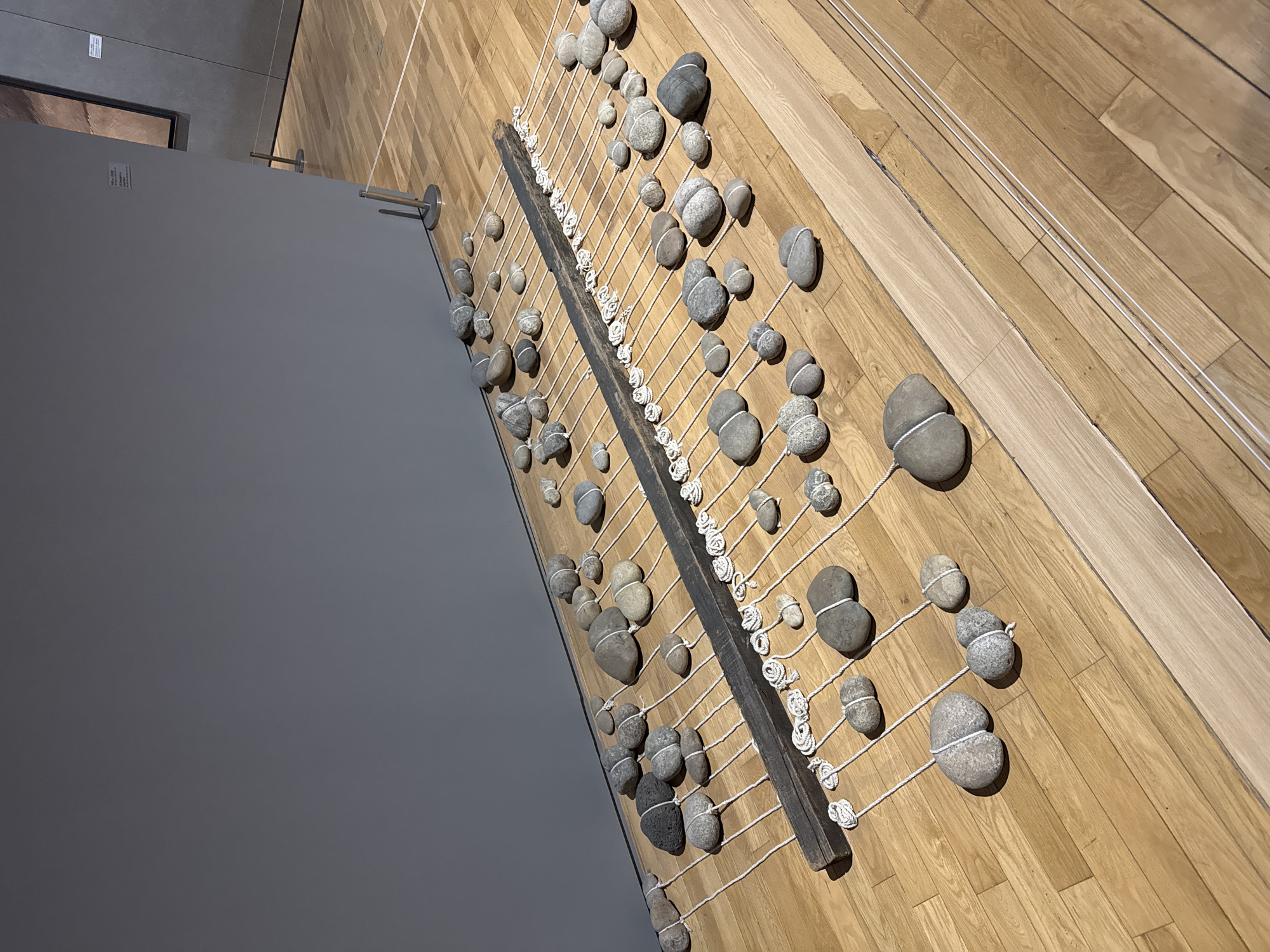 The image shows an art installation on a wooden floor, possibly inside a building. The installation consists of numerous rocks or stones, each tied together in pairs with white twine. These pairs of rocks are suspended by more white twine from a dark, rectangular wooden frame. The twine is knotted at the frame. The artwork is positioned at an angle to the viewer. In the background, a gray wall is visible. The location is likely inside an art gallery or museum. The city of Seoul, South Korea is not discernable from the provided visual information.