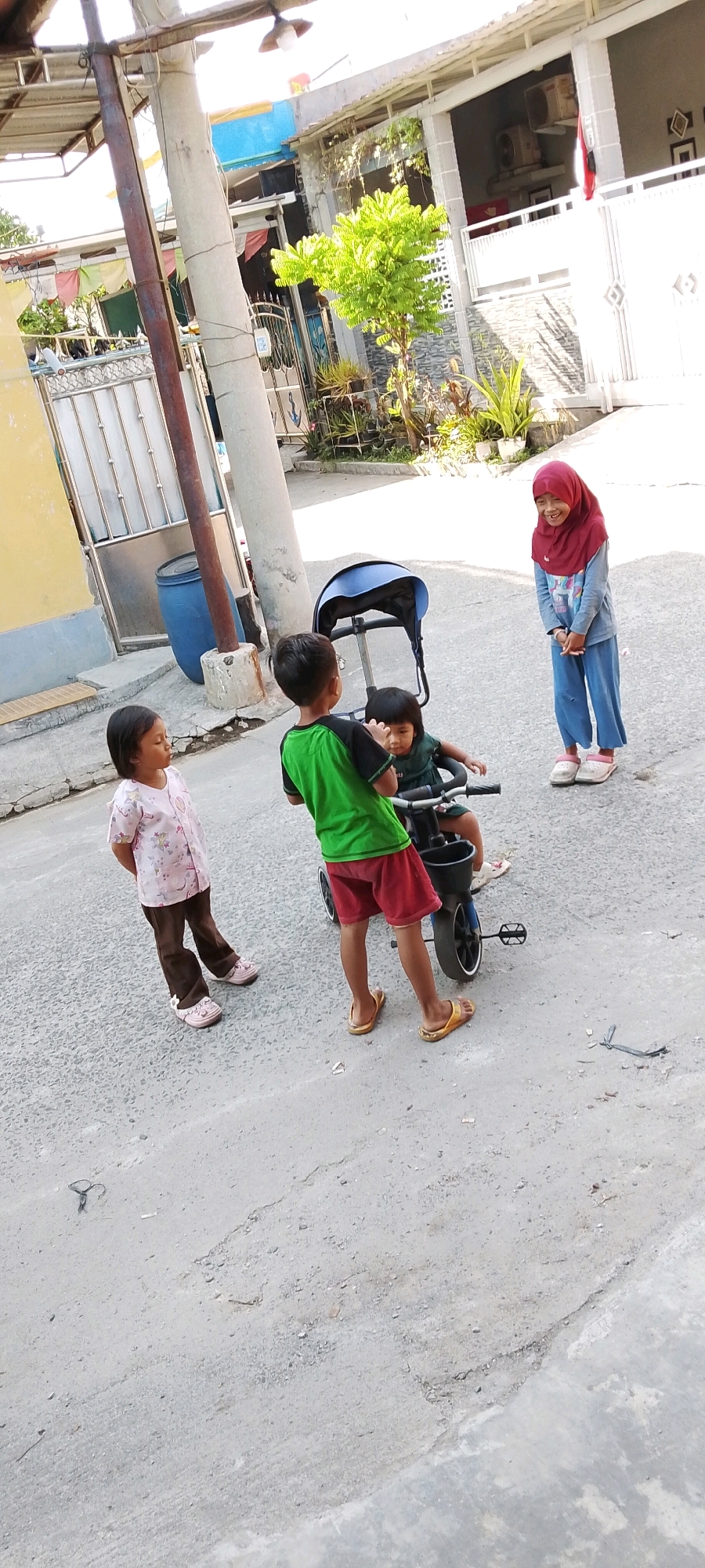 In Satriamekar, Indonesia, four young children are interacting on a paved street. A boy in a green shirt and red shorts is standing behind a blue stroller-type tricycle, holding onto its handlebars. A younger child, wearing a dark green dress, sits inside the tricycle. To the left of the tricycle, a girl in a pink patterned shirt and brown pants stands facing the tricycle. On the right side of the scene, a girl wearing a red hijab and a blue shirt with a graphic print stands facing the other children, with her hands clasped in front of her. The street is paved with a textured grey surface and appears to be cracked in places. In the background, there is a yellow wall on the left, a metal gate, a blue plastic barrel, and a building with a white facade and grey accents. The lighting suggests it is daytime, with shadows cast on the ground.