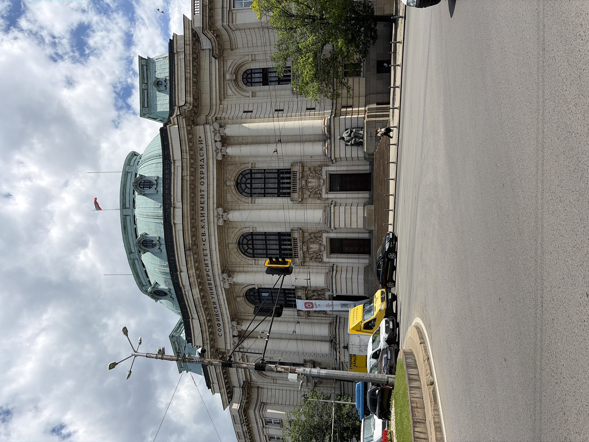 This is a full, eye-level exterior shot of the Sofia University Rectorate building in Sofia, Bulgaria, on a cloudy day. The building is a grand neoclassical structure with a large, green dome and intricate architectural details, including columns and arched windows. The inscription "СОФИЙСКИ УНИВЕРСИТЕТ СВ. КЛИМЕНТ ОХРИДСКИ" is visible above the entrance. A Bulgarian flag flies on a pole atop the dome. In front of the building, a traffic light is illuminated yellow. Several cars are visible on the street, including a yellow delivery van. A statue of a seated man is positioned on a pedestal to the right of the main entrance, and two people are walking in front of the steps leading to the entrance. A large green tree stands to the right of the building. The foreground shows a street intersection with asphalt paving.