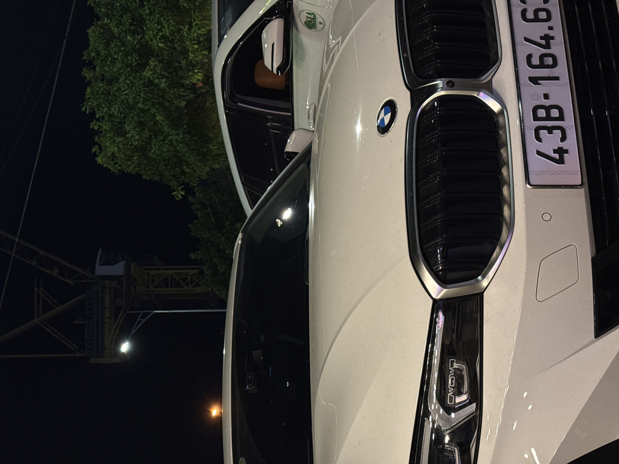 The image depicts a close-up view of the front of a white BMW car, parked at night in the city of Đà Nẵng, Vietnam. In the background, a construction crane is visible, along with trees and a street light. The car's license plate reads "43B-164.63". Another white car is partially visible in the background, with its interior showing tan leather seats. The scene appears to be on a street or parking area within the city.