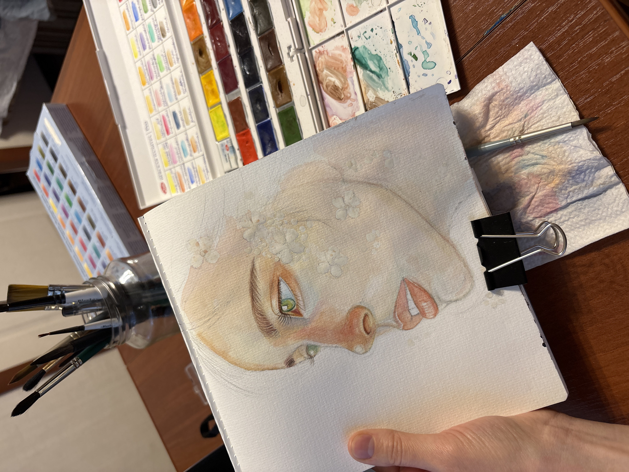The image depicts an art setup on a dark wooden table. A hand holds a watercolor sketchbook. The sketchbook displays an unfinished watercolor painting of a face in profile, adorned with small white flowers. The face has a pale complexion with delicate features; the eye is a vibrant green. To the left of the sketchbook is a glass jar filled with various paintbrushes. Behind the sketchbook are three watercolor palettes. The first palette is a large, open box with rows of watercolor pans. The second is also an open palette with mixed paints in various colors. The third is smaller with individual paints. To the right of the sketchbook sits a crumpled paper towel with paint smudges and a paintbrush resting on top. The background is blurred, suggesting a close-up shot.
There is no indication that the location is in the city of Izhevsk, Russia.
