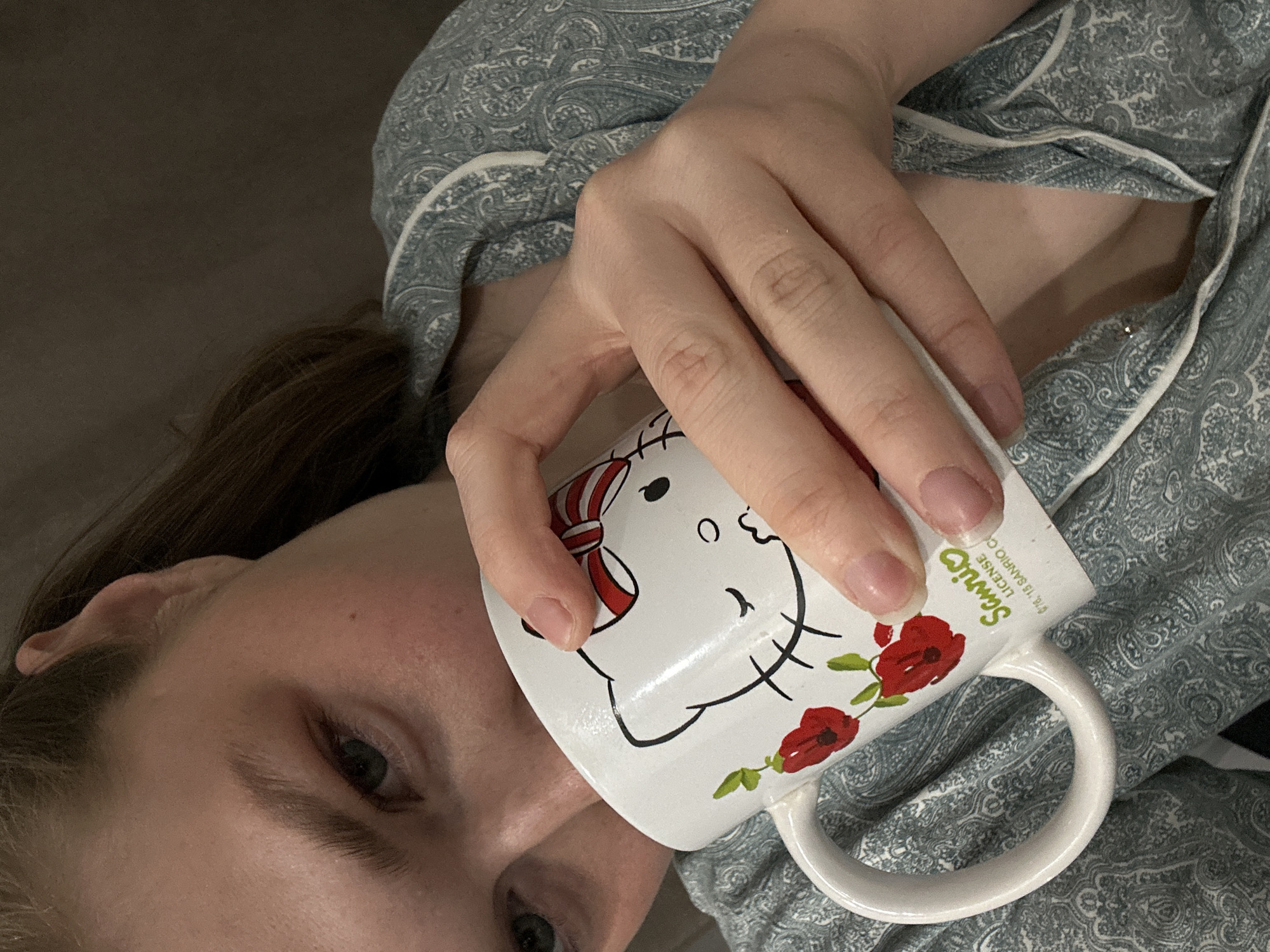 A woman is holding a white mug with a Hello Kitty design, featuring a red bow and floral accents. She is wearing a light blue and white patterned pajama top with white trim. The background is a grey surface, possibly a couch or bed. The location is not discernible from the image.