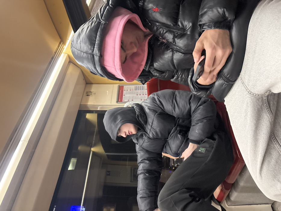 Two individuals are visible inside what appears to be a train or bus. One person is seated, wearing a black puffer jacket with a pink hood underneath. They appear to be sleeping. Another individual beside them is also wearing a black puffer jacket and grey sweatpants. Another person is seated across the aisle in a black jacket and sweatpants, facing forward. The interior consists of beige walls, a window, and informational signage.